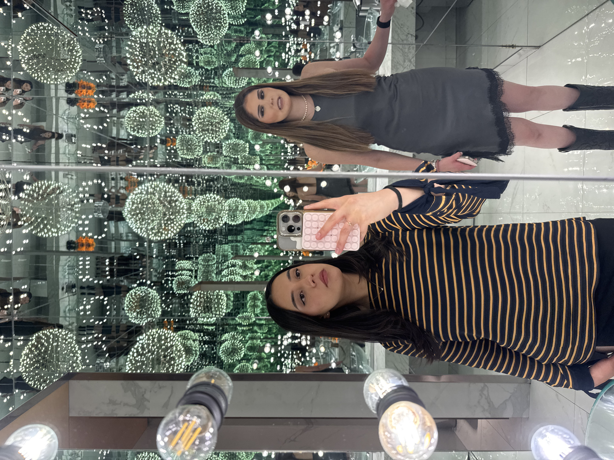 Two young women are posing in a mirror room with walls lined with small, spherical, white lights. The walls are made of gray marble. In the foreground, there are some lightbulbs. The room gives the illusion of infinite space due to the reflections. The location is in the city of Puebla, Mexico. One woman is taking a selfie using a smartphone. She is wearing a black and yellow striped long-sleeved shirt. The other woman is standing behind her and wearing a gray dress and black boots.