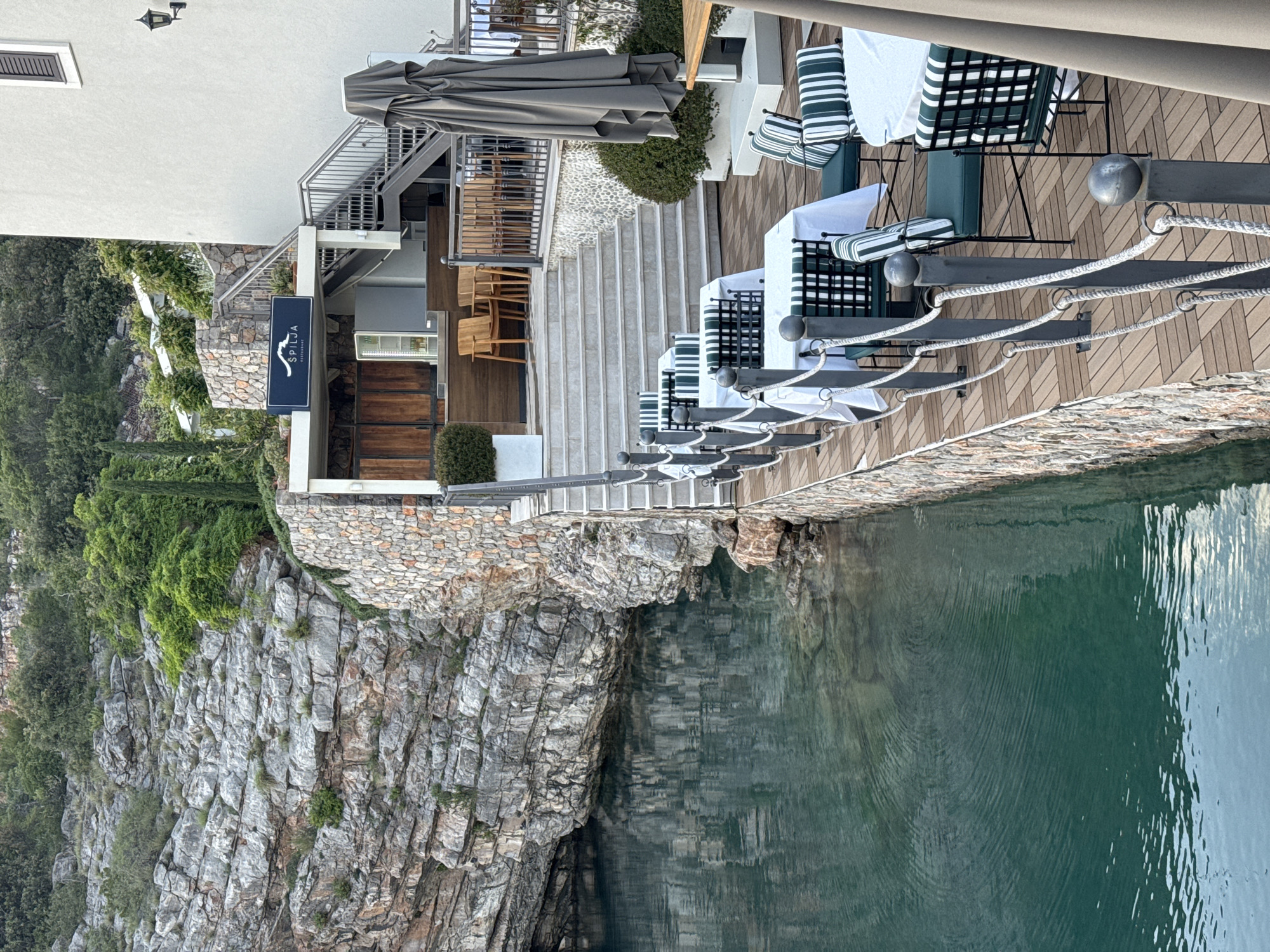 The image depicts a waterfront scene in Radovići, Montenegro, featuring a restaurant ("Spilja") built on a rocky cliffside overlooking the Adriatic Sea. The restaurant has a modern design with a stone facade blending into the natural rock formations. There is an outdoor dining area with tables, chairs, and umbrellas set on a wooden deck that extends over the water. A stone staircase leads from the restaurant down to the sea. The water appears calm and clear with reflections of the cliffside and restaurant. The vegetation covers the cliff. The image appears to be taken during the day, with adequate sunlight illuminating the scene.