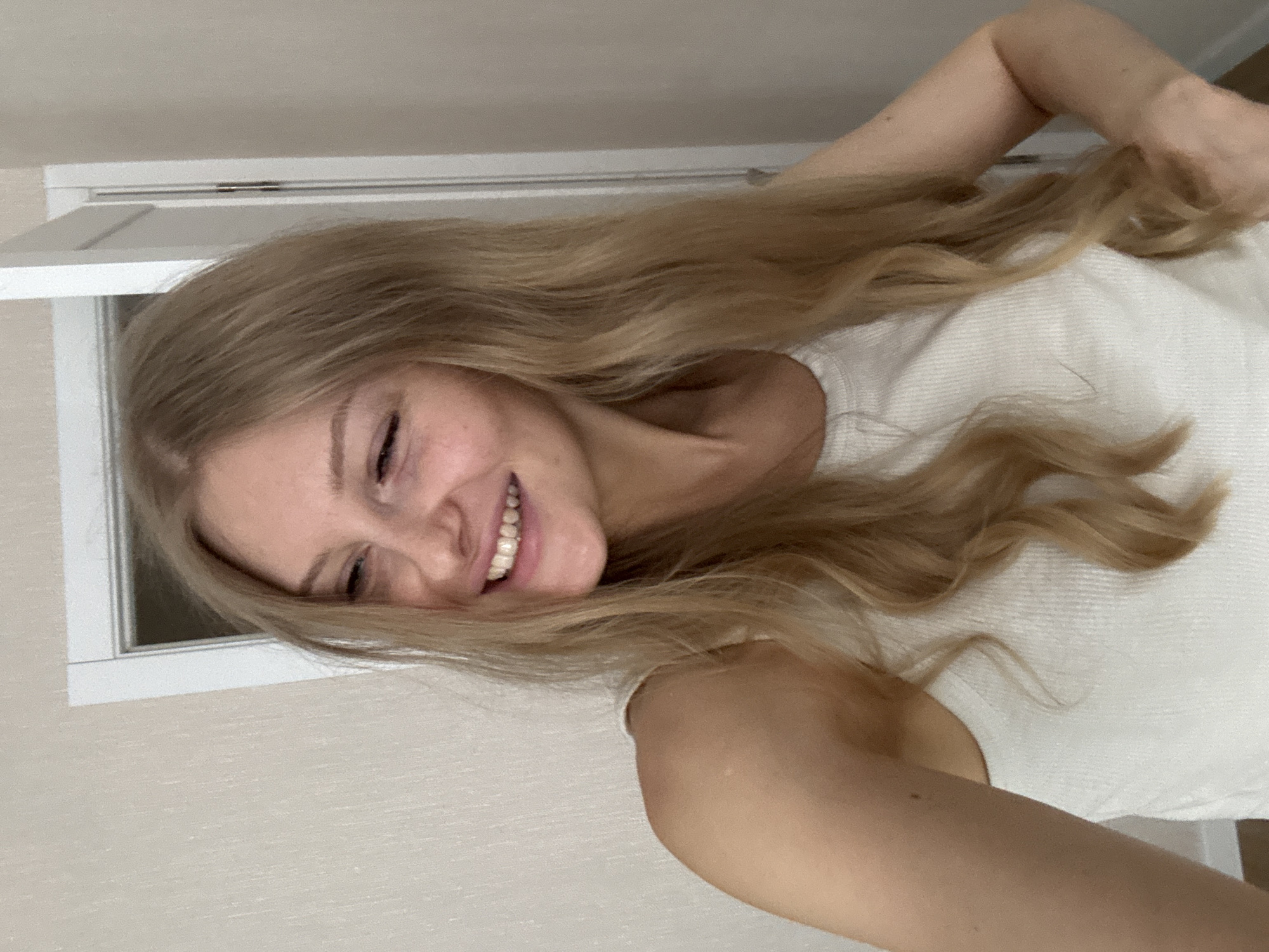 A young woman with long, blonde, wavy hair is smiling at the camera. She is wearing a white tank top. A window is visible in the background. The location is potentially a residential interior, possibly in Perm, Russia.
