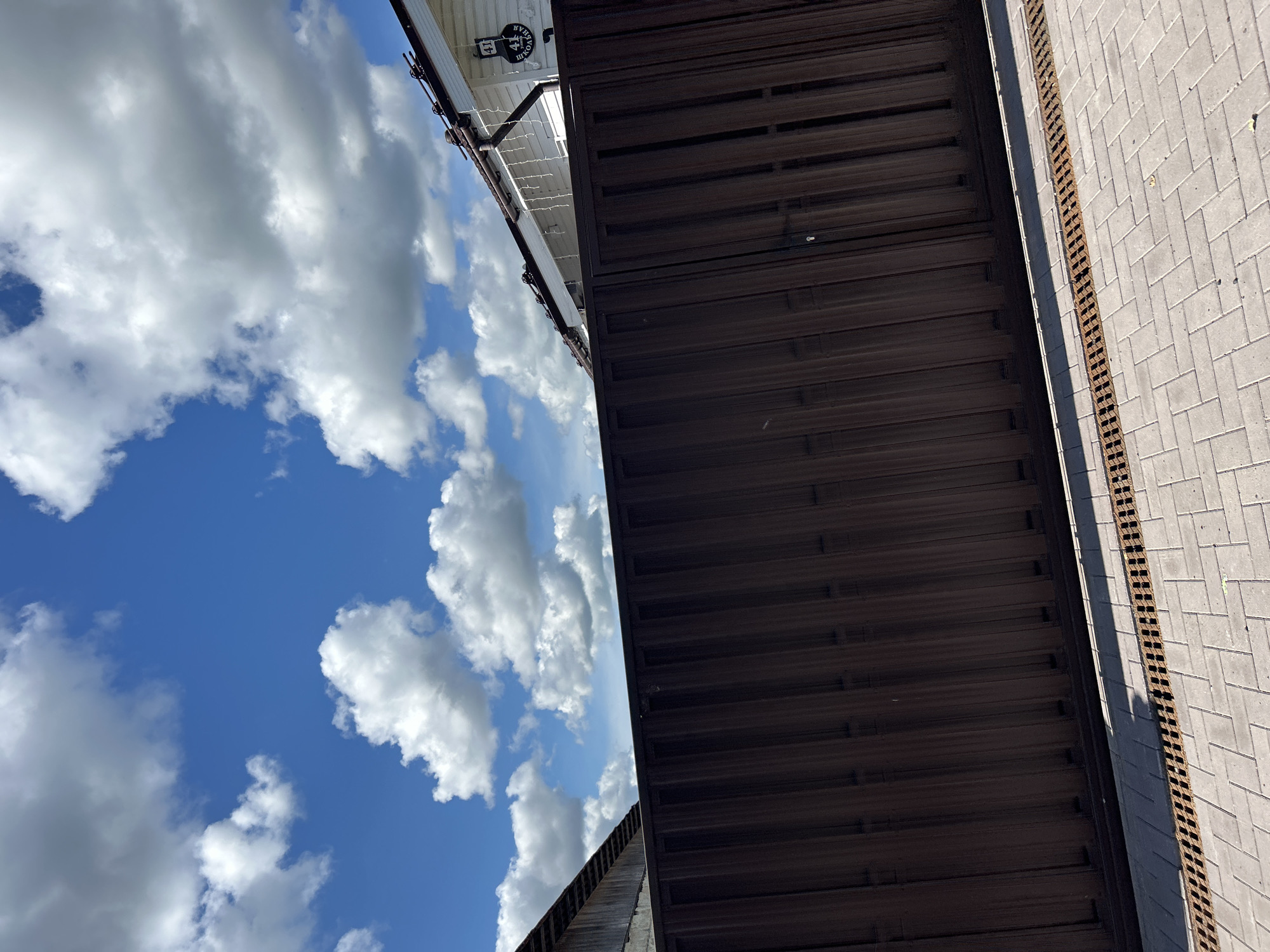 The image depicts a scene outdoors in Kalitsino, Russia, under a blue sky with scattered white clouds. In the foreground, a dark brown metal fence runs horizontally across the frame. The fence appears to be a sliding gate or a section of a larger fence, with vertical ribbed panels. A paved walkway made of light-colored interlocking bricks lies in front of the fence. A narrow metal grate, likely a drainage channel, runs along the base of the fence, separating it from the brick paving.
Behind the fence, a white building with a dark roof is visible. The building has a prominent address sign with the number "41" and text that appears to be in Cyrillic, possibly indicating a street name. Icicle-like decorations hang from the roofline of the building. To the left of the white building, a portion of another structure, possibly a shed or an older building, is visible with a dark, slanted roof. The overall impression is that of a residential or semi-industrial area in a suburban or rural setting. No people are visible in the image.