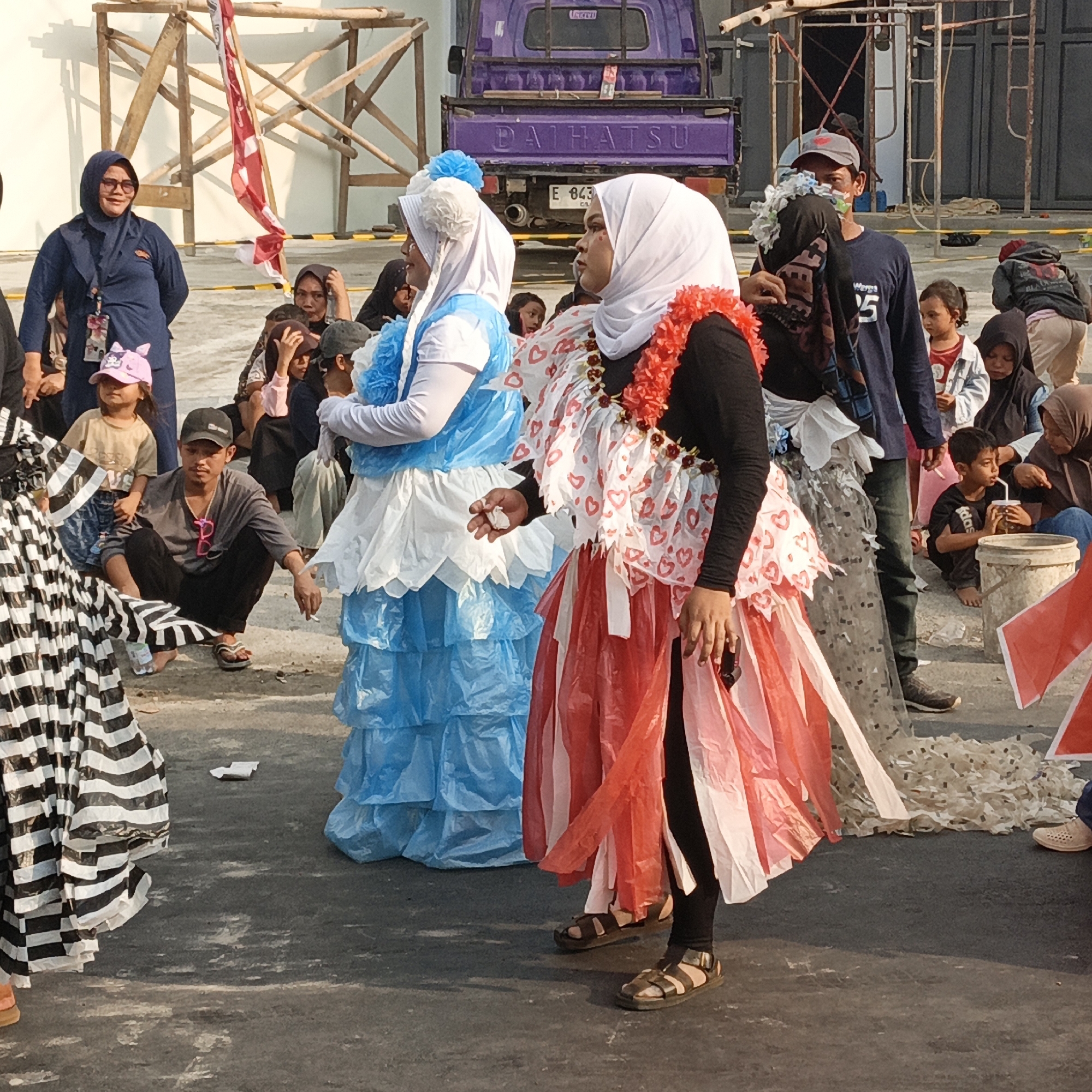 In Randudongkal, Indonesia, a group of people is gathered outdoors, possibly for an event or performance. In the foreground, two women are prominently featured, both wearing creatively designed dresses made from what appears to be plastic material. The woman on the left is wearing a white and blue layered dress, and the woman on the right is in a dress with red and white heart patterns, adorned with a red flower lei. Behind them, other individuals are visible, including children and adults, some sitting and some standing. In the background, a purple truck with "DAIHATSU" written on it is parked, and scaffolding structures are present, suggesting construction or an outdoor event setup. The ground is paved, and a small amount of debris is scattered around. The lighting indicates it is daytime.
