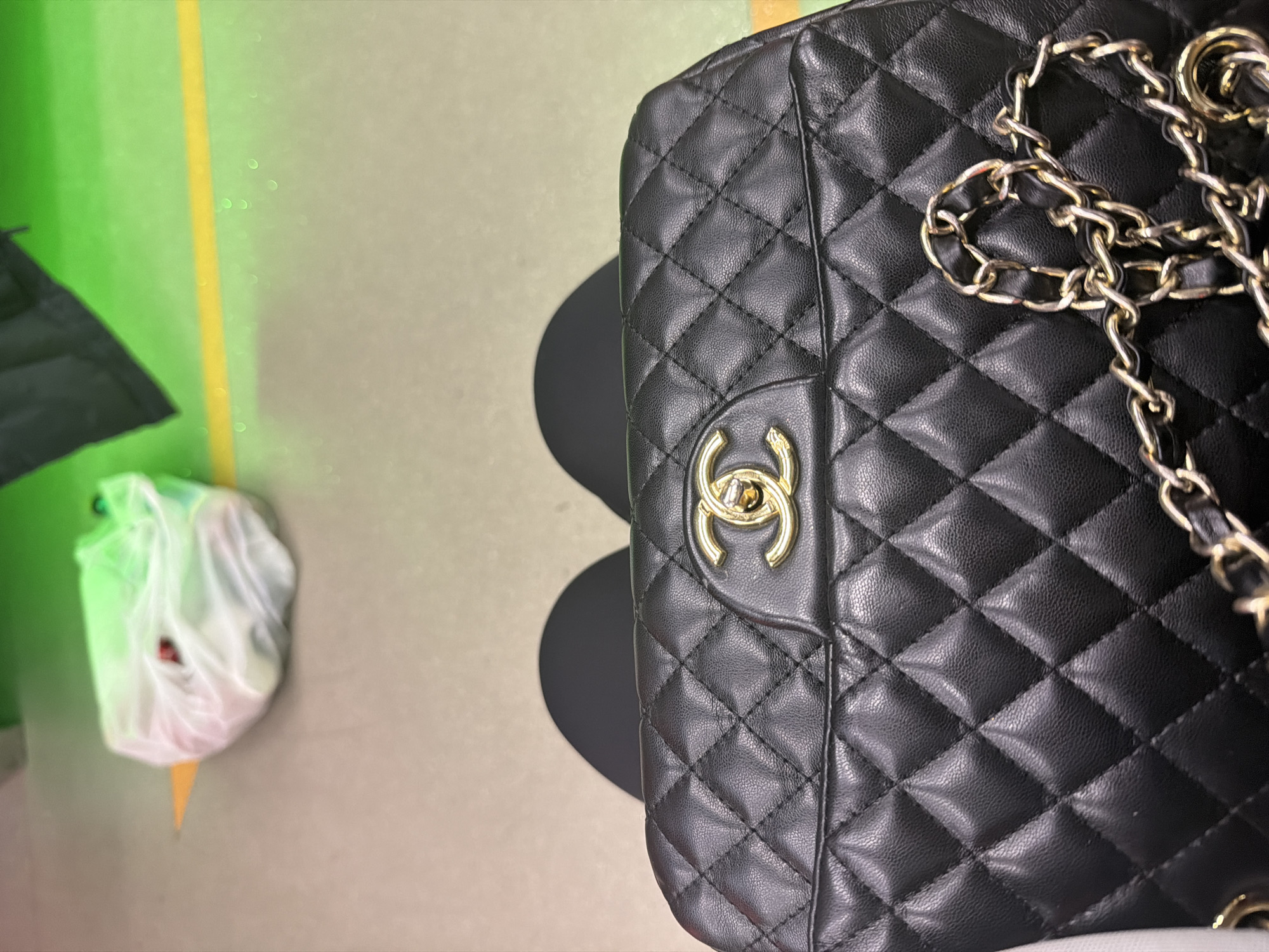 An overhead shot shows a black, quilted Chanel bag resting on someone's lap. The bag features a gold-toned interlocking CC logo closure and a interwoven chain strap. In the background, a white plastic bag is visible on a light gray and yellow-lined floor, possibly in a public area such as an airport terminal or transit station. The environment appears to be indoors with bright, overhead lighting casting reflections on the floor.