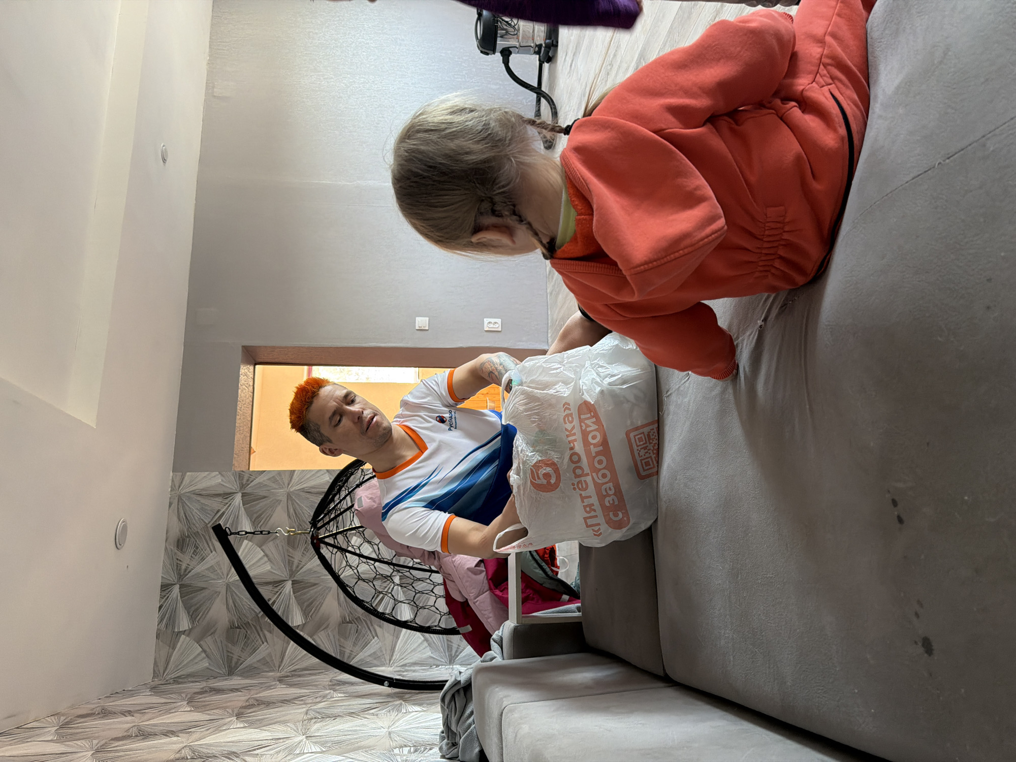 The scene depicts a person with bright orange hair, wearing a white and blue athletic shirt, seated in a hanging chair within a room. A child with pigtails, dressed in an orange jumpsuit, leans over the edge of a gray couch. The person is handing a plastic shopping bag labeled "Пятёрочка" (Pyaterochka), a Russian grocery store chain, towards the child. The environment includes a geometric patterned wall and floor, suggesting a residential interior. This photo was taken in the city of Chaikovsky, Russia.