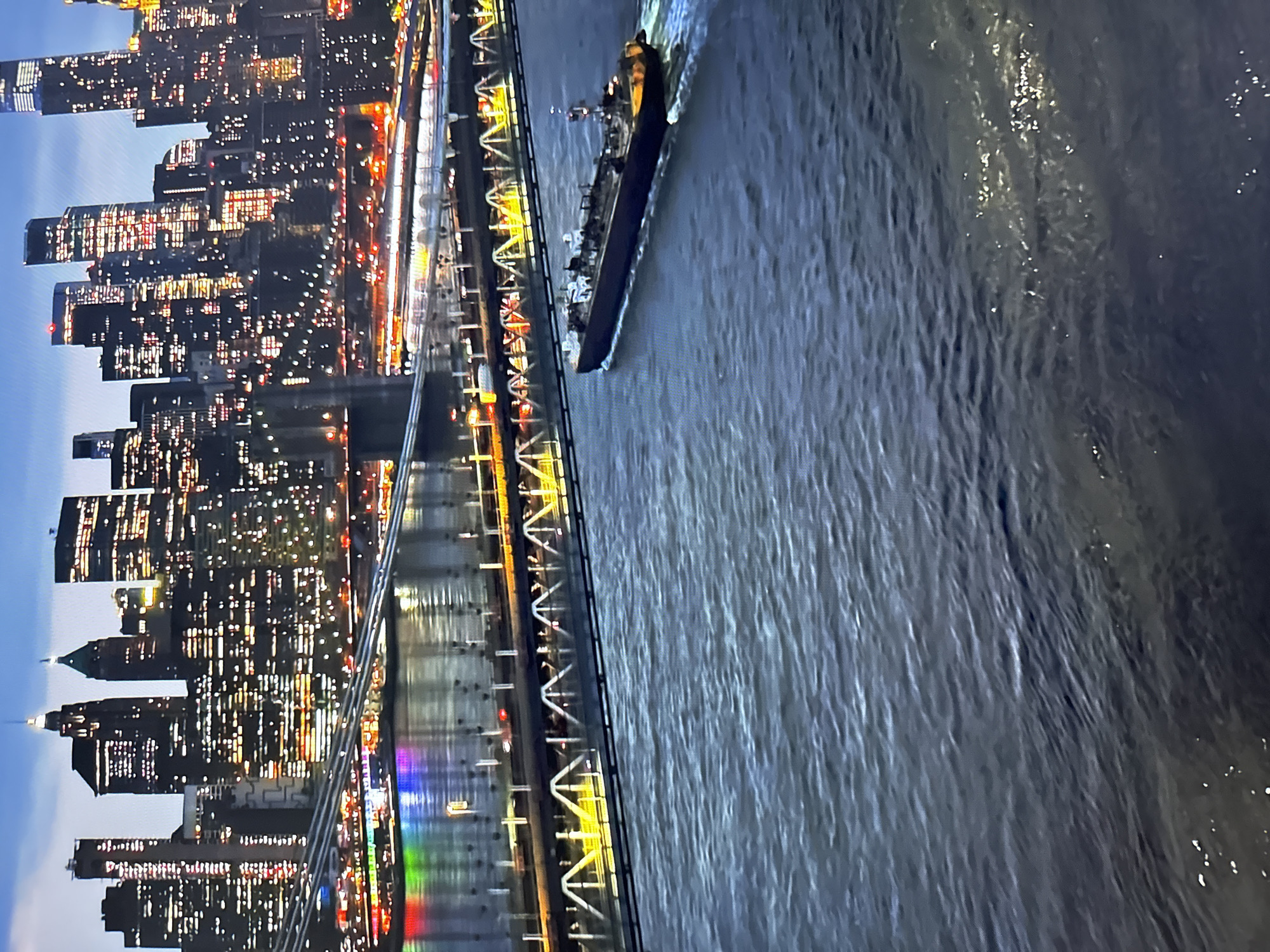The image shows a wide shot of the New York City skyline at dusk. The city is illuminated by countless lights from the skyscrapers, and the Brooklyn Bridge is visible in the foreground. A barge is sailing on the water in front of the bridge. The water is dark and choppy, with ripples reflecting the lights from the city. The sky is a deep blue, with a few faint clouds. There are no people visible in the image. The scene is bustling with urban activity, with the city lights creating a vibrant and dynamic atmosphere.