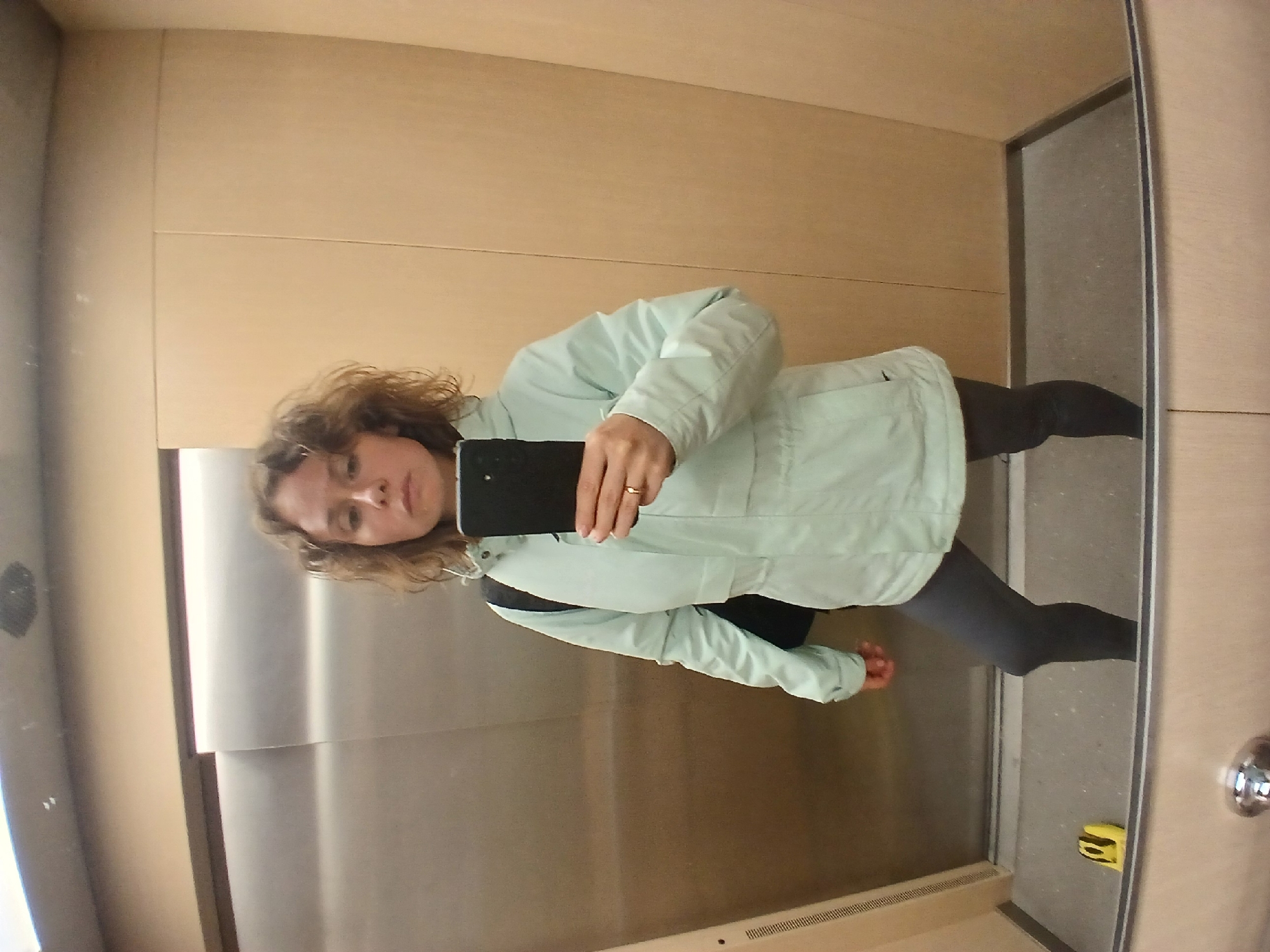 A woman with curly brown hair stands in an elevator and takes a selfie in the mirror. She is wearing a light green hooded jacket and dark grey leggings. She has a black backpack slung over her left shoulder and is holding a black smartphone in her right hand, which is positioned to capture the reflection. A gold ring is visible on her right ring finger. The elevator walls are a combination of brushed metal and light wood paneling. The floor of the elevator is grey and speckled. A small yellow object with black markings is visible on the floor near the elevator doors. The mirror reflects the lower half of the elevator, including the floor and a portion of the door frame. The scene is forensic and matter-of-fact, depicting a common urban environment.