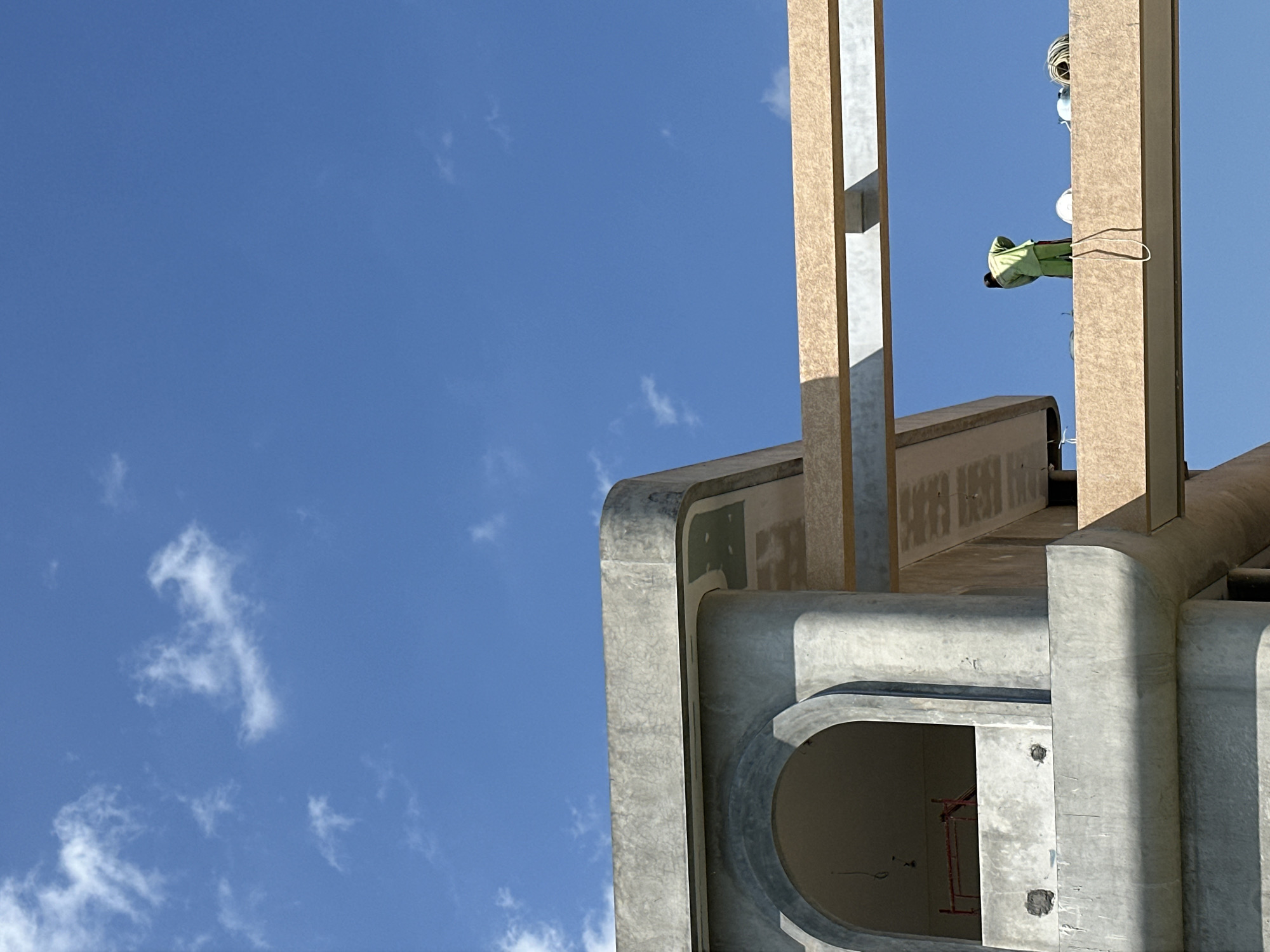 In North Kuta, Indonesia, a construction site features a partially built concrete structure against a bright blue sky dotted with scattered white clouds. The building exhibits rounded architectural elements and a large arched opening. Two horizontal beams, appearing to be made of engineered wood, span across the foreground, casting shadows. On the upper beam, a person wearing a green uniform is visible, facing away from the camera, engaged in activity. Several white hard hats and a coil of rope are also present on the beam. The scene captures a moment of construction work under daylight.