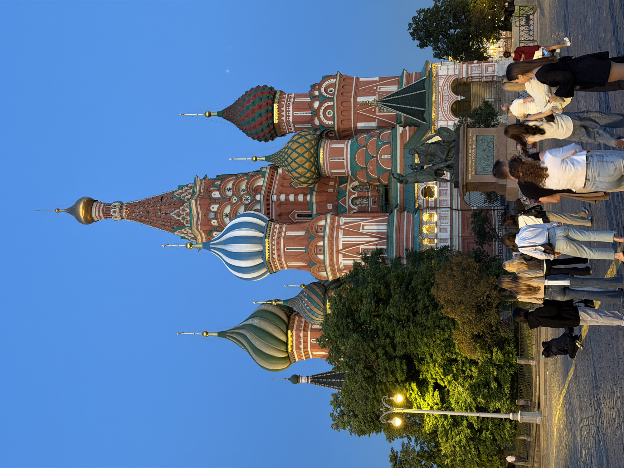 This image captures St. Basil's Cathedral and the Minin and Pozharsky Monument in Red Square, Moscow, Russia, during twilight or early evening. The sky is a deep, clear blue.
St. Basil's Cathedral, a prominent architectural landmark, dominates the background. Its red brick walls are adorned with intricate patterns, and its multiple onion domes feature vibrant colors and designs, including stripes of blue and white, and geometric patterns in green, red, and gold. The central spire is topped with a golden dome. Subtle lighting illuminates portions of the cathedral's lower sections.
In the mid-ground, positioned in front of the cathedral, is the Minin and Pozharsky Monument, a bronze sculptural group depicting two historical figures on a pedestal.
The foreground features a group of approximately ten individuals, mostly young adults, standing on cobblestone pavement. Most are viewed from behind or the side and are facing towards the cathedral, suggesting they are tourists or observers of the monument and cathedral. Their attire is casual. One person on the left is crouched low to the ground. Another person, further back on the right, is walking away.
To the left, a tall, white streetlight with two yellow-glowing lamps illuminates a dense cluster of green trees. A metal fence separates the paved area from the trees. The ground is paved with irregularly shaped cobblestones.