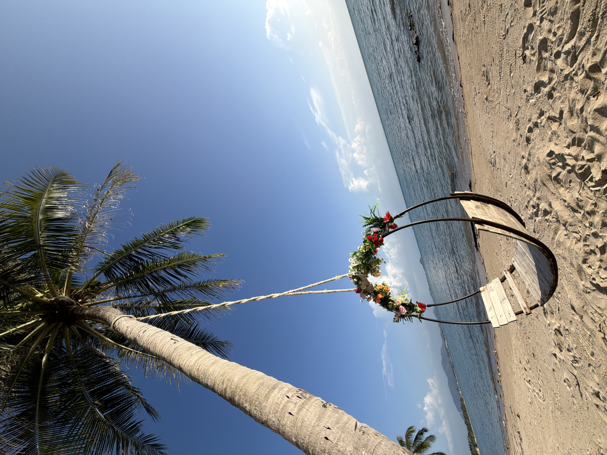 An empty wooden swing decorated with colorful flowers hangs from a thick rope tied to a palm tree on a sandy beach in the Ko Samui District of Thailand. The swing is positioned to face the calm blue ocean, with gentle waves lapping at the shore. In the background, a distant coastline with mountains is visible under a clear blue sky with scattered white clouds. The sand on the beach has footprints and is illuminated by the bright sunlight, casting subtle shadows. The scene evokes a sense of peaceful tranquility.