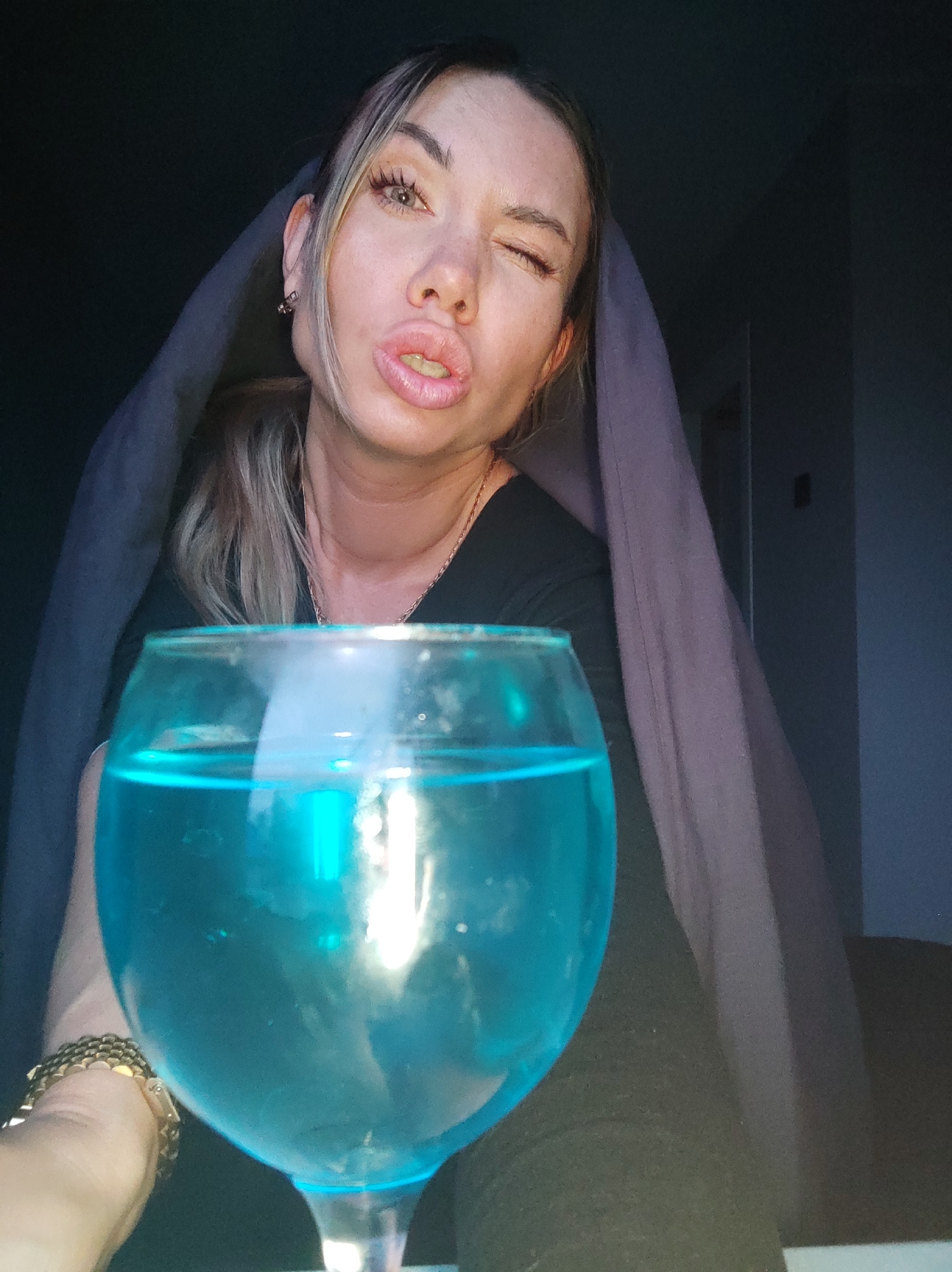A woman is posing with a glass of blue liquid. She is wearing a dark top, and a blanket is draped over her head. She is winking and pursing her lips. A gold bracelet is visible on her wrist. The setting appears to be indoors, in a dimly lit room. There are no readily identifiable landmarks to confirm the location as Votkinsk, Russia.
