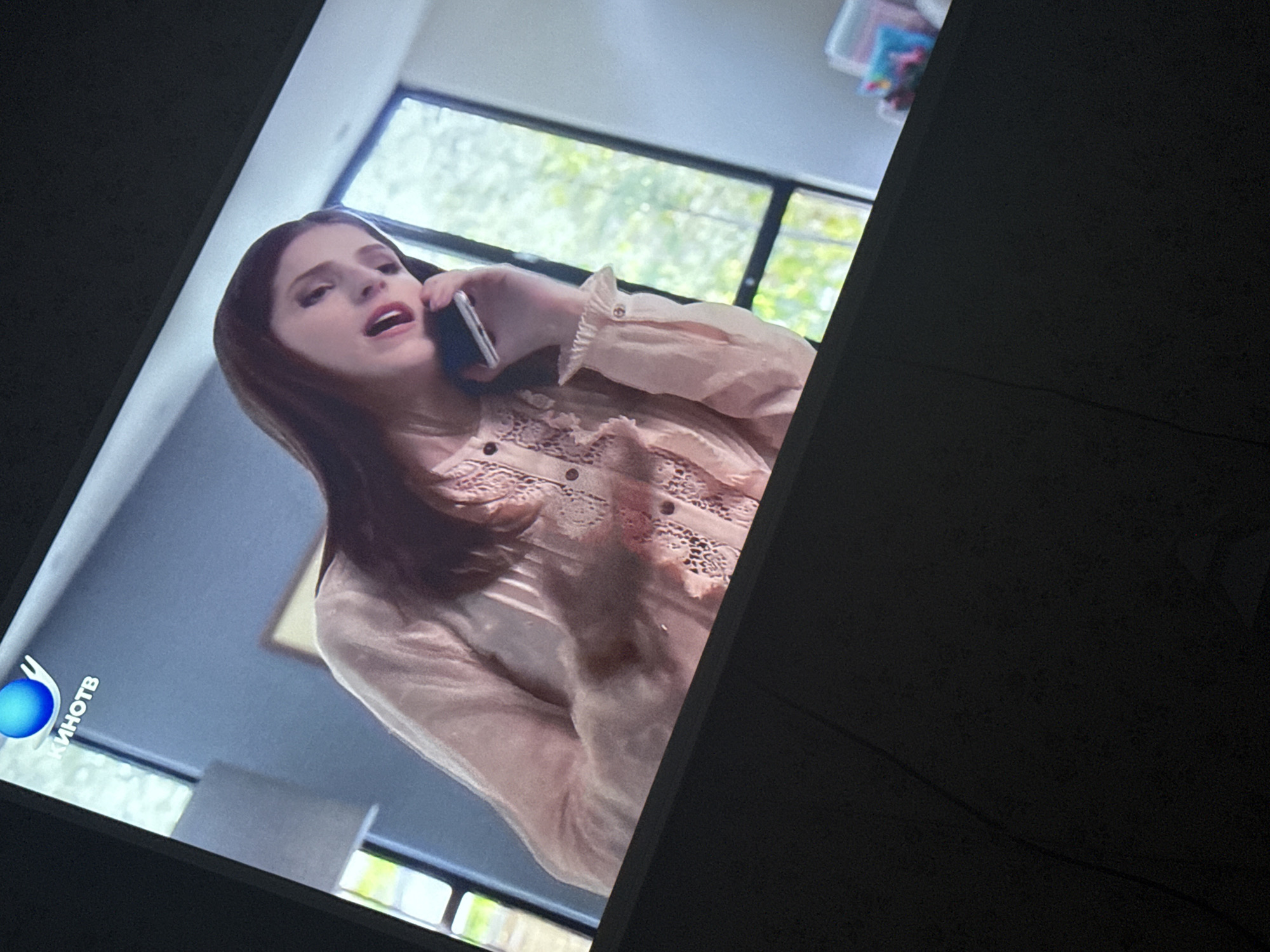 The image shows a screen displaying a woman with brown hair, wearing a light-colored blouse with lace detail, and holding a smartphone to her ear, as if engaged in a phone call. The background appears to be an interior setting with a window revealing trees. A logo of "КИНОТВ" is visible in the lower-left corner of the screen. There's insufficient information to determine the exact location in Mytishchi, Russia, or any specific actions/interactions taking place.