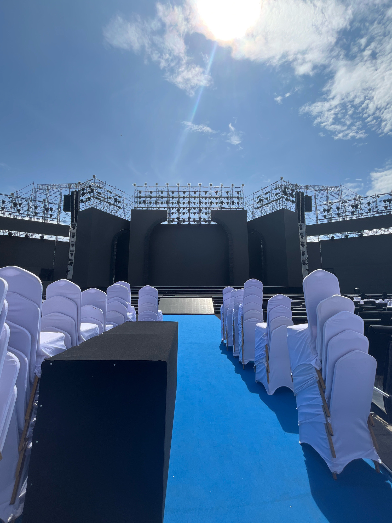 An outdoor stage is set up with rows of white-covered chairs facing it. The stage is large and dark, with multiple tiers and a large screen at the back. Above the stage, there is a complex structure of scaffolding with lights and speakers. The sky is blue with some scattered clouds and the sun is shining brightly. A small airplane is visible in the distance. The scene appears to be set up for an event, possibly a concert or festival, in Nha Trang, Vietnam. The overall impression is one of anticipation and preparation for a large-scale gathering.