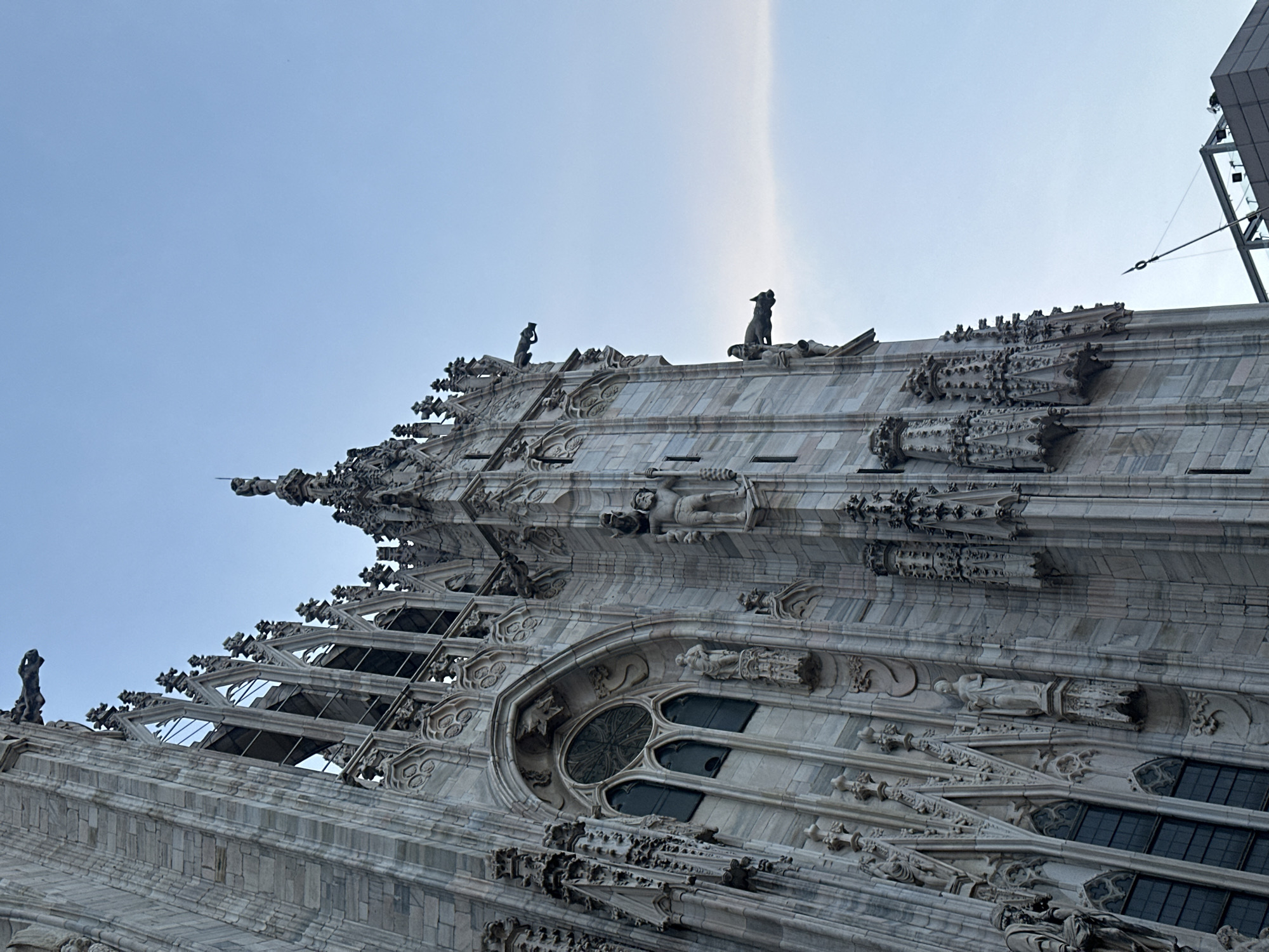 The image shows a close-up, low-angle view of the intricate stone facade of the Milan Cathedral (Duomo di Milano) in Milan, Italy, against a clear blue sky. The architecture is characterized by gothic spires, ornate carvings, statues, and gargoyles. A prominent circular stained-glass window with geometric patterns is visible on the left side of the frame. The stonework is light-colored, appearing to be marble, and exhibits a weathered texture. The overall impression is one of grandeur and historical significance.