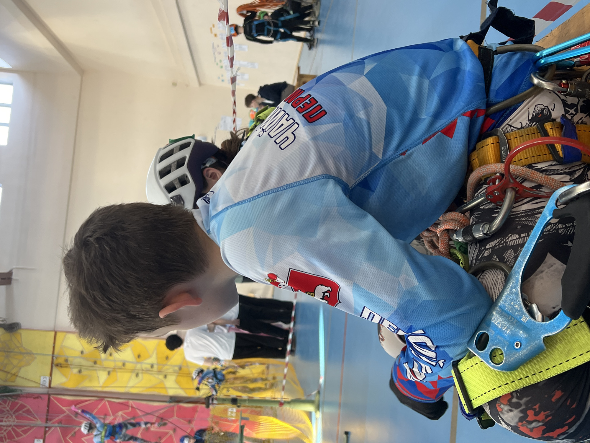 Here is a description of the image:
The image depicts a young person with short brown hair, wearing a white climbing helmet and a blue and white athletic shirt with red lettering that reads "Пермь", which translates to "Perm", and a coat of arms associated with the city and region of Perm, Russia. The climber is outfitted with a climbing harness and various pieces of climbing gear including ropes, carabiners, and a belay device. The climber is looking down towards a colorful indoor climbing wall with other climbers visible in the background. The photograph was likely taken in Berezniki, Russia.