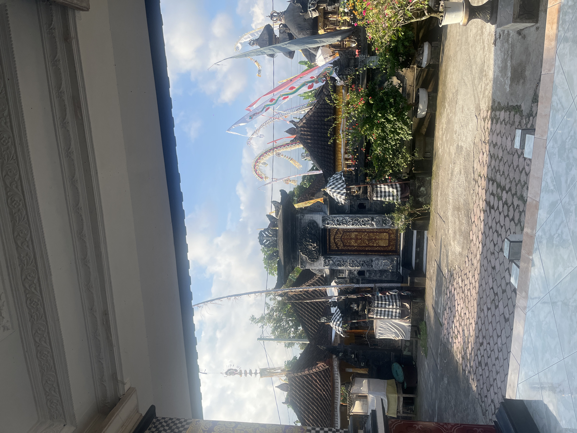 The image shows a courtyard in front of traditional Balinese buildings, likely a temple or a residential compound, in Mendoyo, Indonesia. The architecture features dark, intricately carved stone structures with ornate golden doors. Several buildings have tiered, dark-tiled roofs. Colorful flags, some with dragon-like motifs, are flying in the breeze. Decorative banners and tall poles with offerings are also visible. Green foliage and flowering plants add to the scene. The ground is paved with interlocking stones in some areas and concrete in others, with sunlight casting shadows. The sky is blue with scattered clouds. The view is framed by a white, ornate ceiling structure, suggesting the photo was taken from inside a building looking out. There are no people visible in the image.