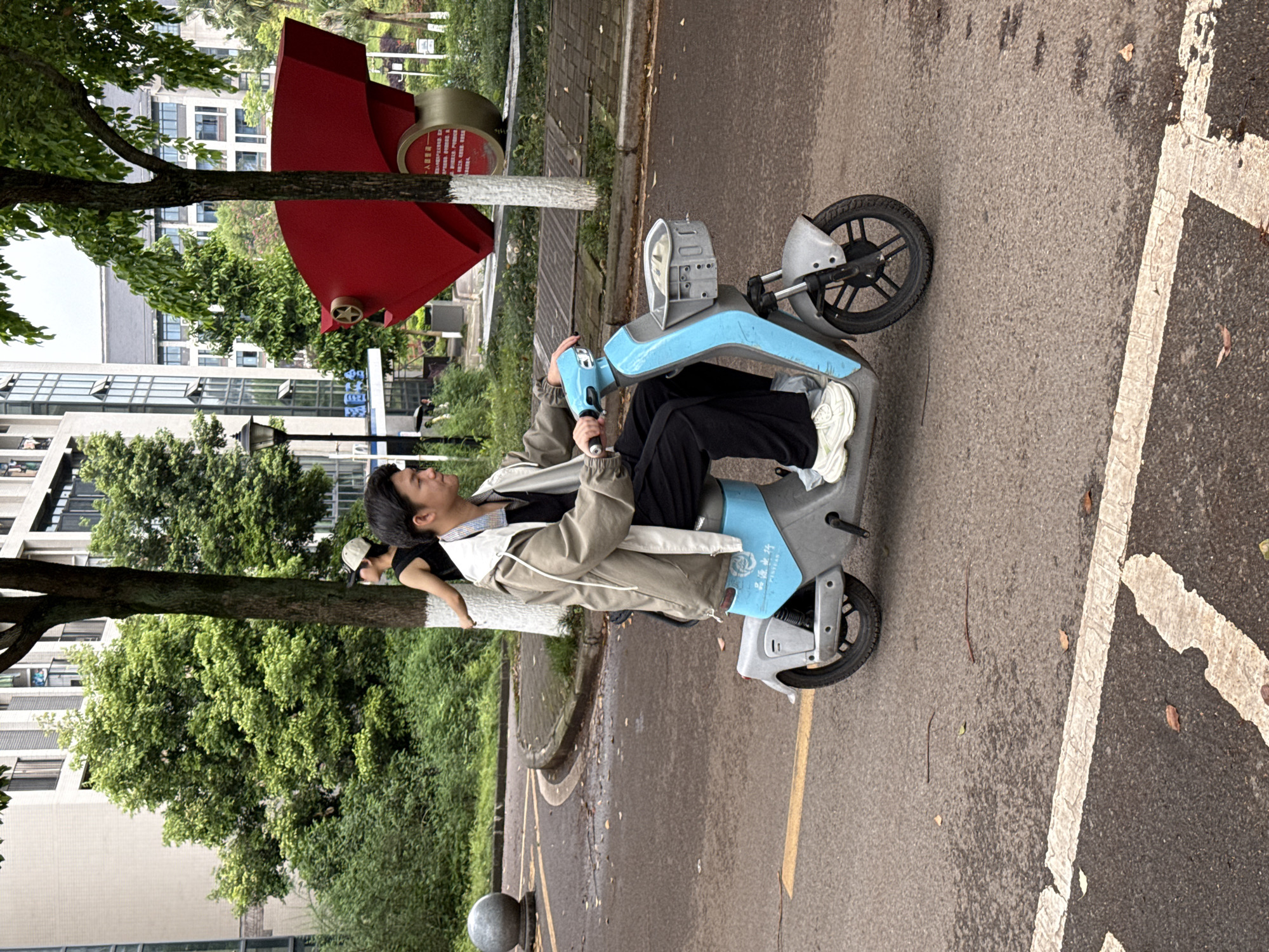 A person wearing a beige jacket, black pants, and white sneakers is sitting on a blue electric scooter on a paved road. A second person, wearing a white baseball cap, is standing beside the scooter. The location is in Jiangjin, China. In the background, there is a red and white cylindrical structure beside a road and trees. Buildings are visible in the distance behind the trees. White painted parking space dividers are on the right side of the image.