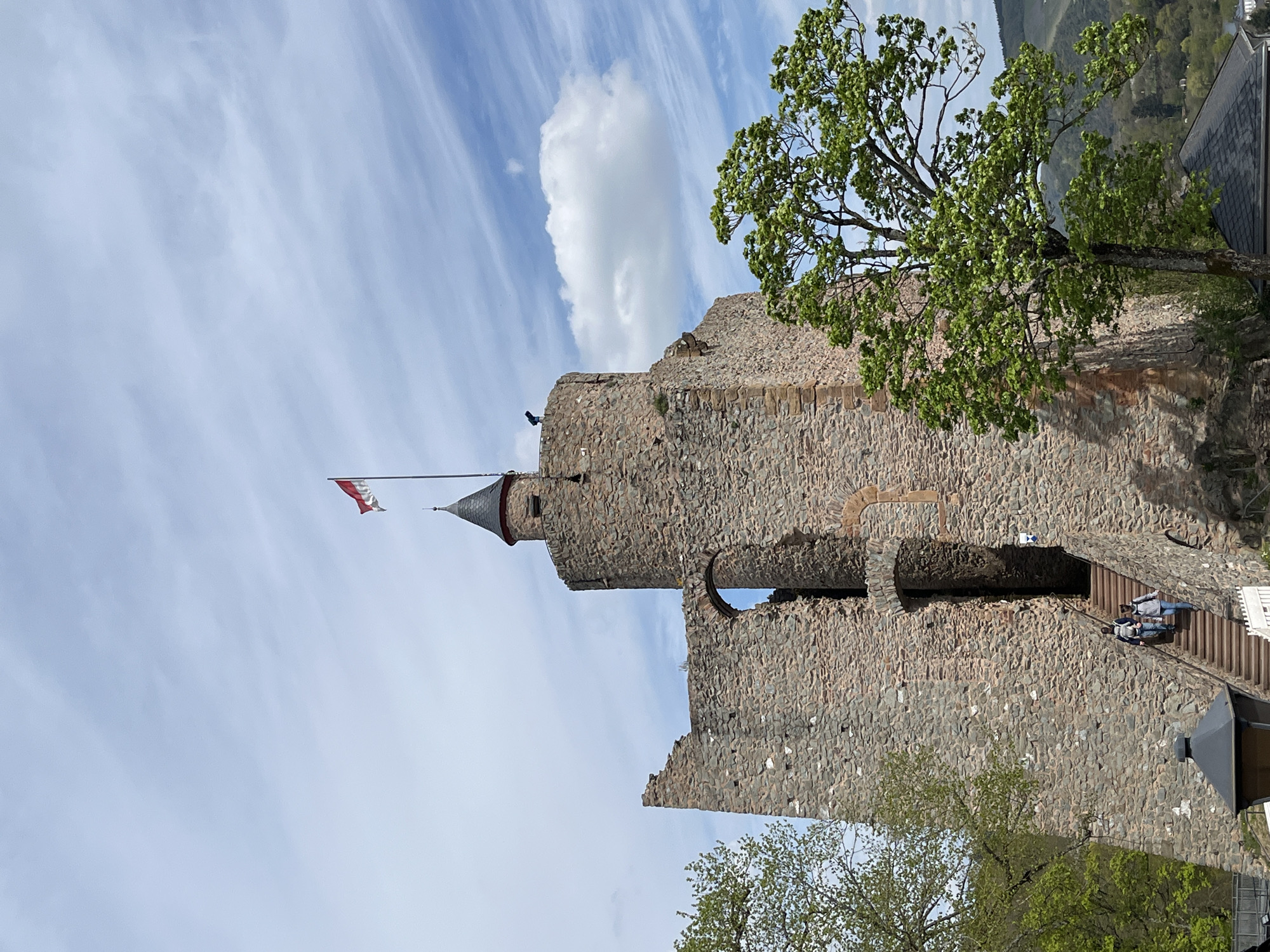 The image shows a partial view of Saarburg Castle in Saarburg, Germany, taken from a lower vantage point. The castle ruins are constructed of grey stone and feature a prominent tower with a conical roof and a red and white flag atop. A stone archway is visible connecting parts of the castle. Adjacent to the castle wall, a wooden staircase leads upwards, with two individuals visible on the steps. Vegetation including a tree with green leaves is present on the right side of the image. The sky is light blue with white clouds.