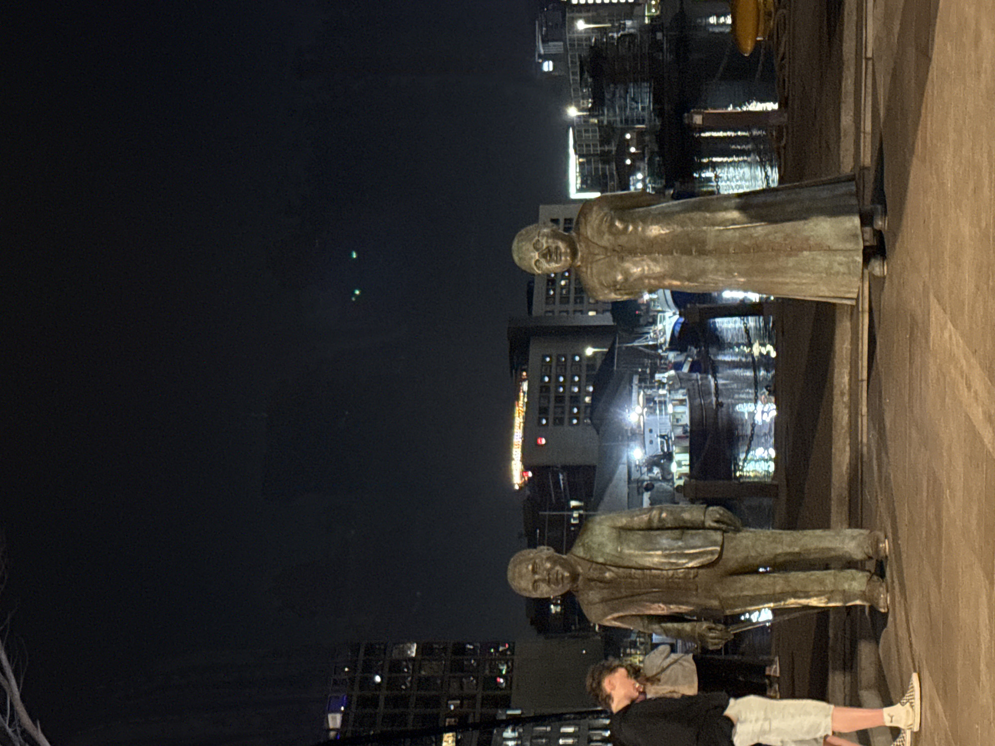 The image shows two bronze statues of a man and a woman, positioned on a stone platform overlooking a waterfront area with buildings and lights reflecting in the water. A person is lying on the platform near the man's statue. The setting appears to be at night in an urban area. While not definitively confirmed, this scene could be at the V&A Waterfront in Cape Town, South Africa, based on the statue style and waterfront setting.
