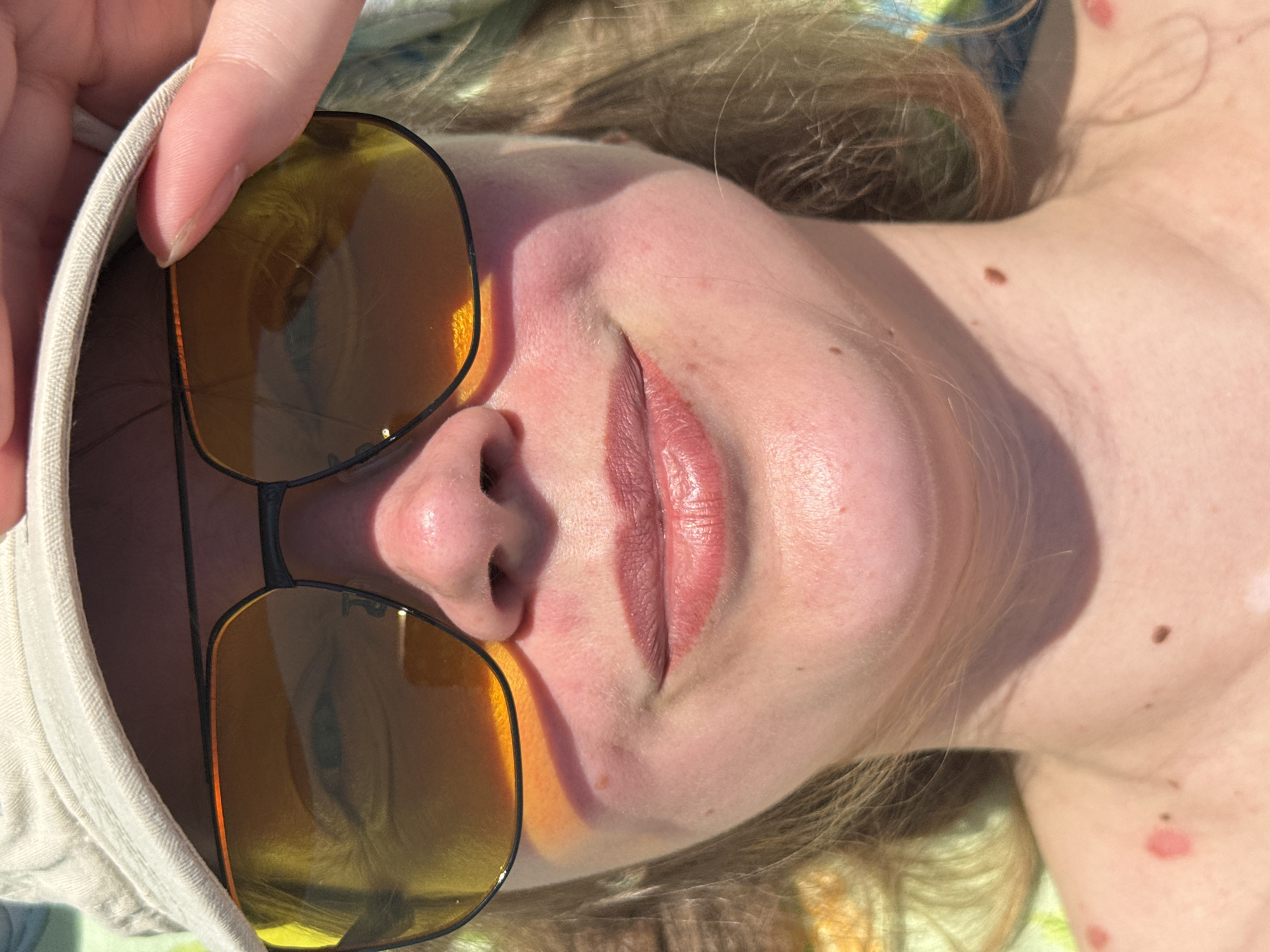 The image is a close-up, slightly angled, portrait of a fair-skinned person wearing sunglasses. The person is lying down, possibly outdoors, in bright sunlight. The sunglasses have gold-tinted lenses and a black frame, held by a hand with a light-colored sleeve on what appears to be a cap. The person's lips are visible, appearing pink and full. There are some moles visible on the person's neck and chest. The background consists of fair hair. The location is not discernible from the image and cannot be determined to be Izhevsk, Russia.