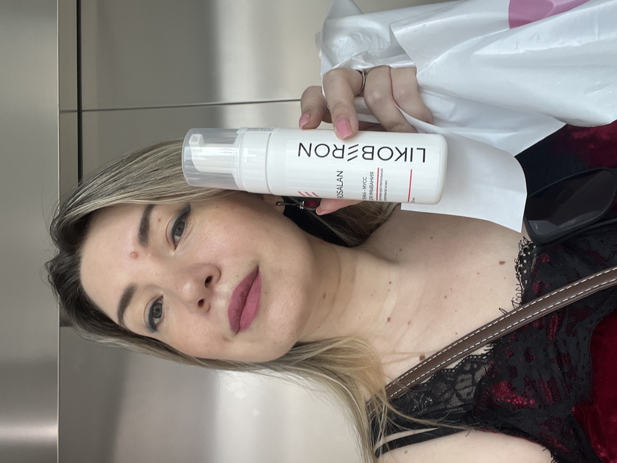 A medium shot captures a woman holding a white bottle of LIKOBERON facial cleanser. She is positioned in front of what appears to be an elevator door, suggesting an indoor setting. The woman has fair skin, blonde hair with highlights, and is wearing dark clothing with lace detailing. The bottle's label is partially visible, displaying Russian text. The background is a metallic surface, typical of an elevator interior.