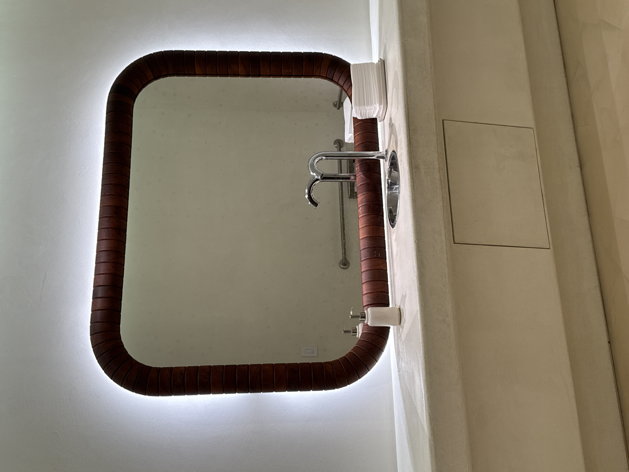 The image shows a bathroom interior. A rectangular mirror with rounded corners is mounted on a wall, featuring a wooden frame and illuminated edges. The mirror reflects a sink, faucet, and soap dispenser. Adjacent to the mirror is a beige wall structure with a rectangular access panel. The scene is located in San Diego, United States of America.