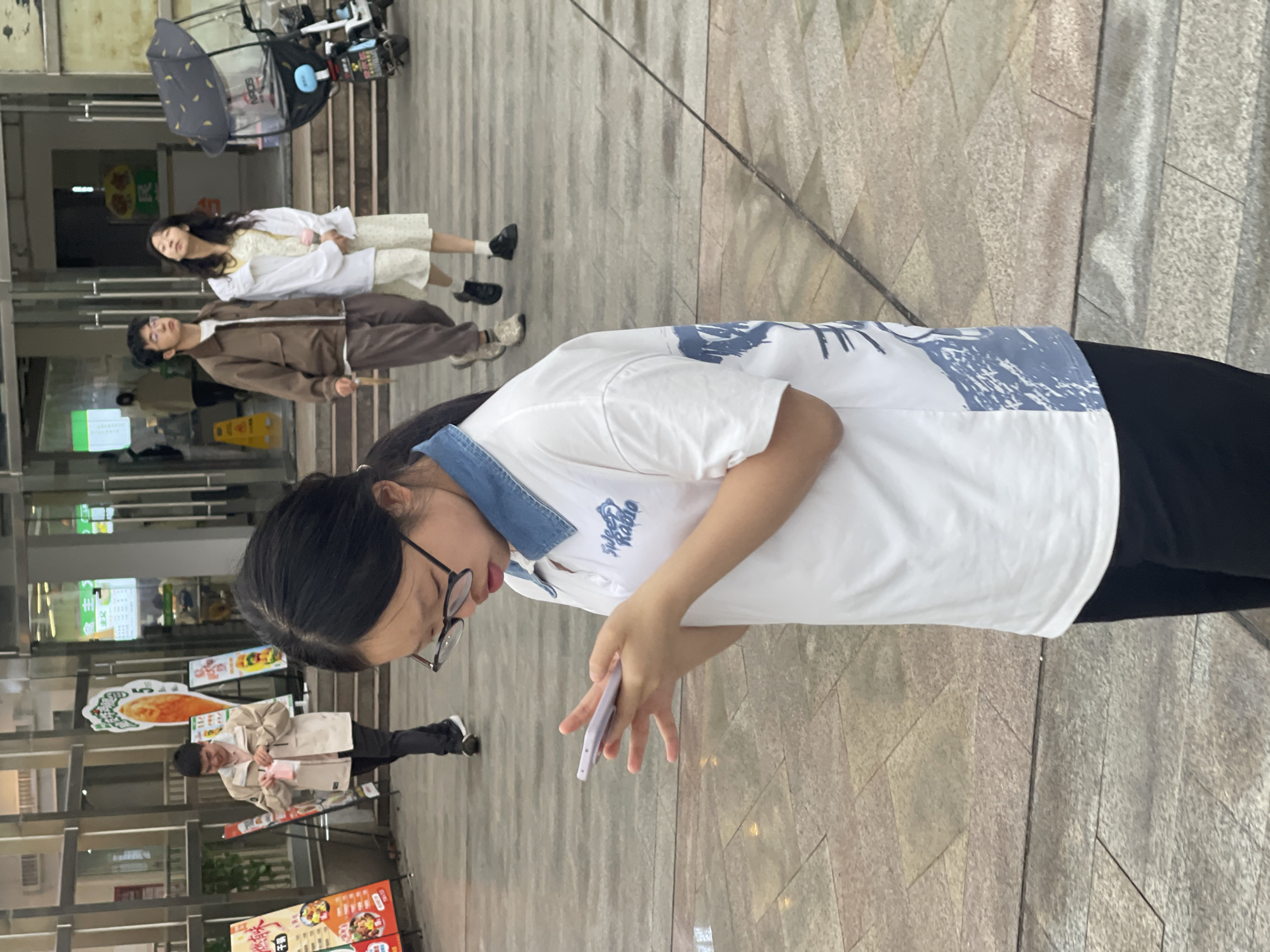 A young woman with dark hair and glasses stands in front of a building in the city of Chongqing High-tech Zone, China. She is wearing a white polo shirt with blue accents and black pants, and she is holding a mobile phone. In the background, two people are lying on the ground in front of an open shop with a motorcycle in the foreground and another person in front of an establishment with food posters displayed.
