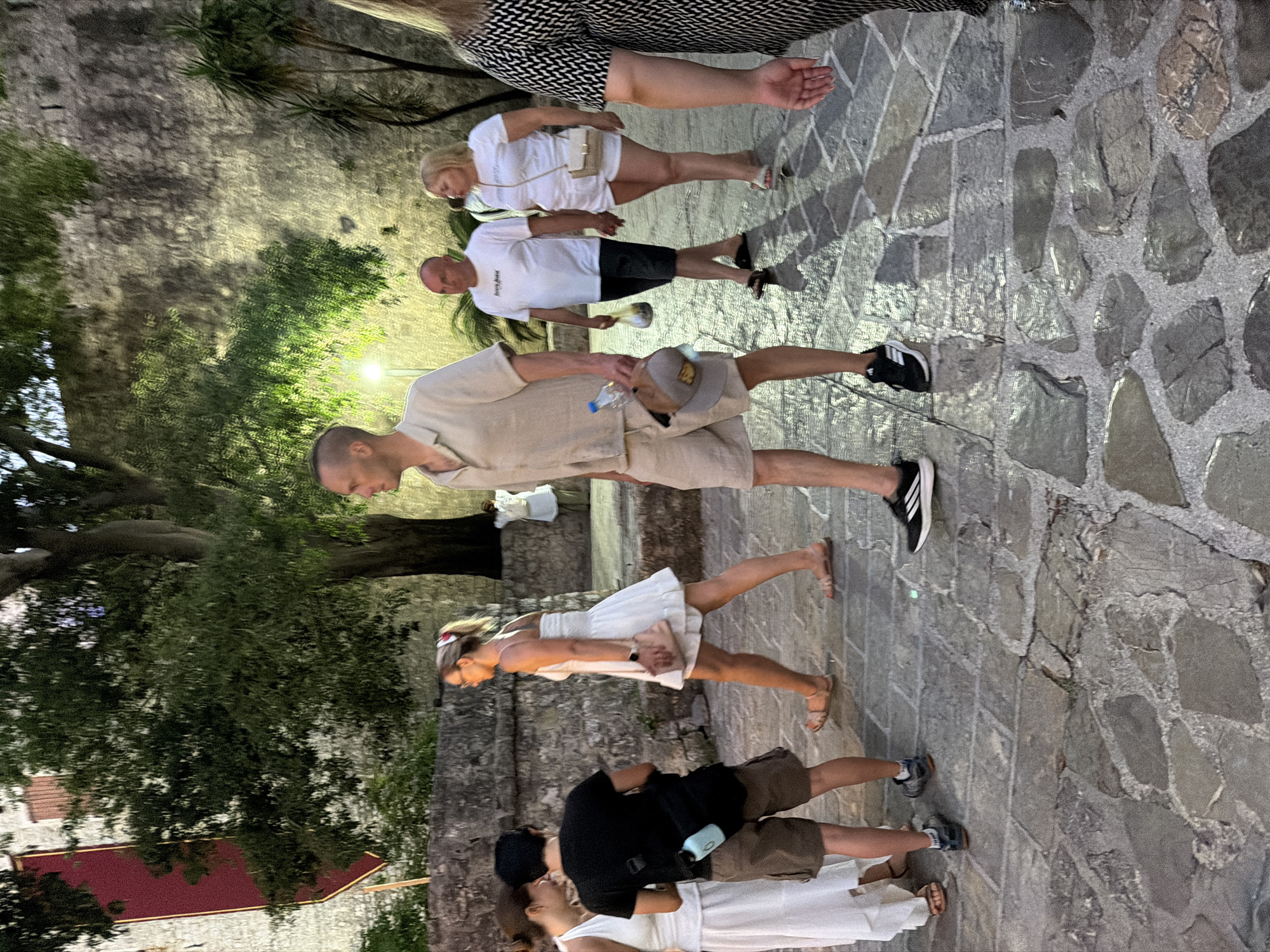 The image shows a group of people walking on a cobblestone street in Budva, Montenegro, with a stone wall in the background. The lighting suggests it is evening or nighttime, with artificial lights illuminating the scene.
In the foreground, a man is walking from left to right, wearing a light-colored short-sleeved shirt and matching shorts. He is holding a water bottle in his right hand and a cap in his left. To his left, a woman in a white dress is also walking to the right, carrying a pink clutch. To the left of her, another woman in a white dress is walking away from the camera, and a man in a black t-shirt and brown shorts is walking alongside her, with a bag slung over his shoulder containing a light blue water bottle.
Further back, a man in a white t-shirt and dark shorts is walking with a woman in a white t-shirt and white shorts. The woman is carrying a small beige bag. Another person, mostly obscured by the woman in the foreground, is wearing a black and white patterned garment and is partially visible on the right side of the frame.
The environment features a textured stone wall behind the people and a large tree with dense foliage. The cobblestone pavement is uneven, with large, irregularly shaped stones.