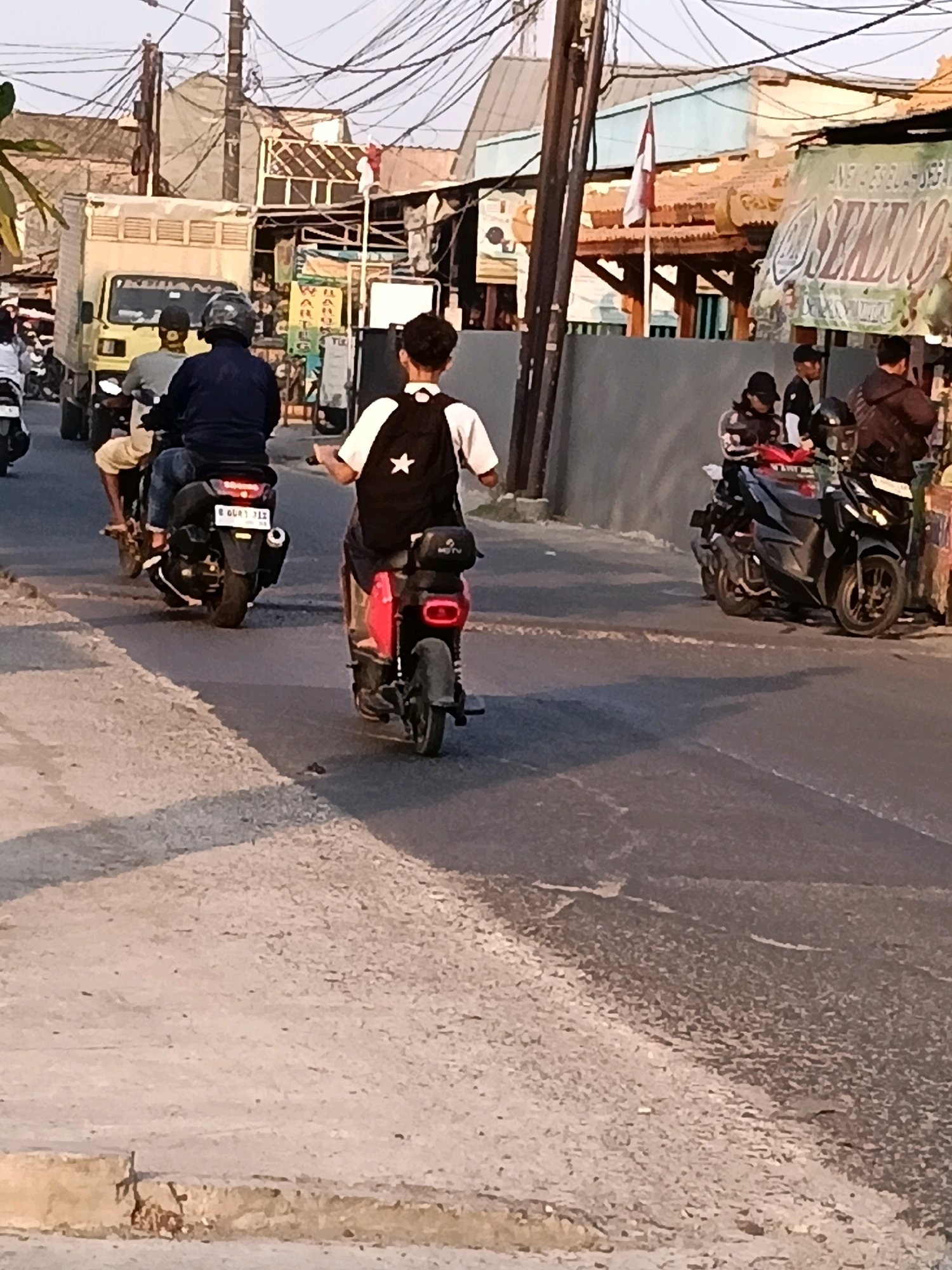 The image depicts a street scene in the city of Belimbing, Indonesia. A person is riding a red scooter from behind, wearing a black vest with a white star and a white shirt. To the left, another person is riding a black scooter. Further to the left, a yellow truck is visible, with two people on a motorcycle in front of it. Several other motorcycles are parked on the right side of the road, with people standing around them. The road is paved, and the surroundings show buildings and a utility pole with numerous wires. The sky is clear, suggesting it is daytime.