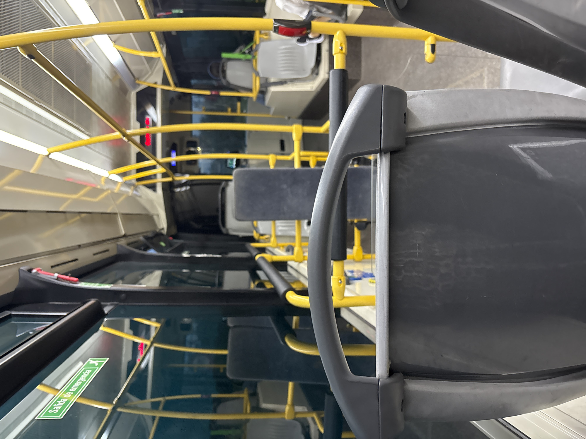 The image shows the interior of a bus, possibly in the city of Córdoba, Spain, as indicated by the emergency exit sign reading "Salida de emergencia". The focus is on the safety features and layout of the bus. Yellow handrails are prominent throughout the vehicle. A grey passenger seat is visible in the foreground, with a handle. The general seating arrangement of the bus is visible in the background, where some seats are equipped with headrests. The image is taken from the front of the bus, with a reflection on the windows. The overall ambiance suggests a standard public transportation environment.