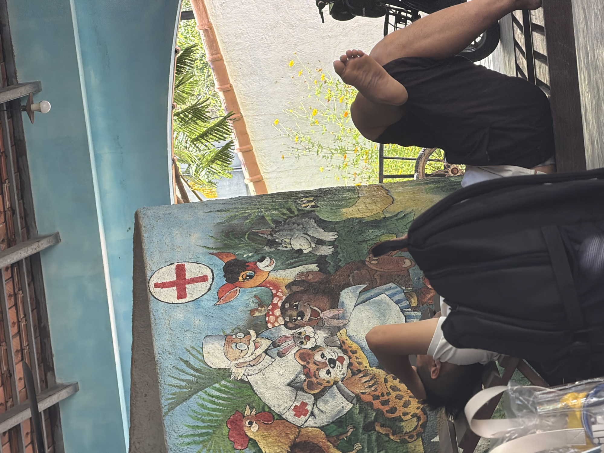 A person is resting on a bench next to a mural. The mural depicts a doctor with various animals, including a chicken, a deer, a cat, a tiger, a rabbit, and a bear. A red cross symbol is visible on the doctor's attire and on a separate emblem. The person is wearing dark shorts and a white t-shirt, with their legs crossed and feet exposed. A black backpack is placed on the bench in front of them. The background shows vegetation, a white wall, and a tiled roof. The setting appears to be an outdoor area, possibly a public space or a recreational area in Tay Nha Trang Ward, Vietnam.