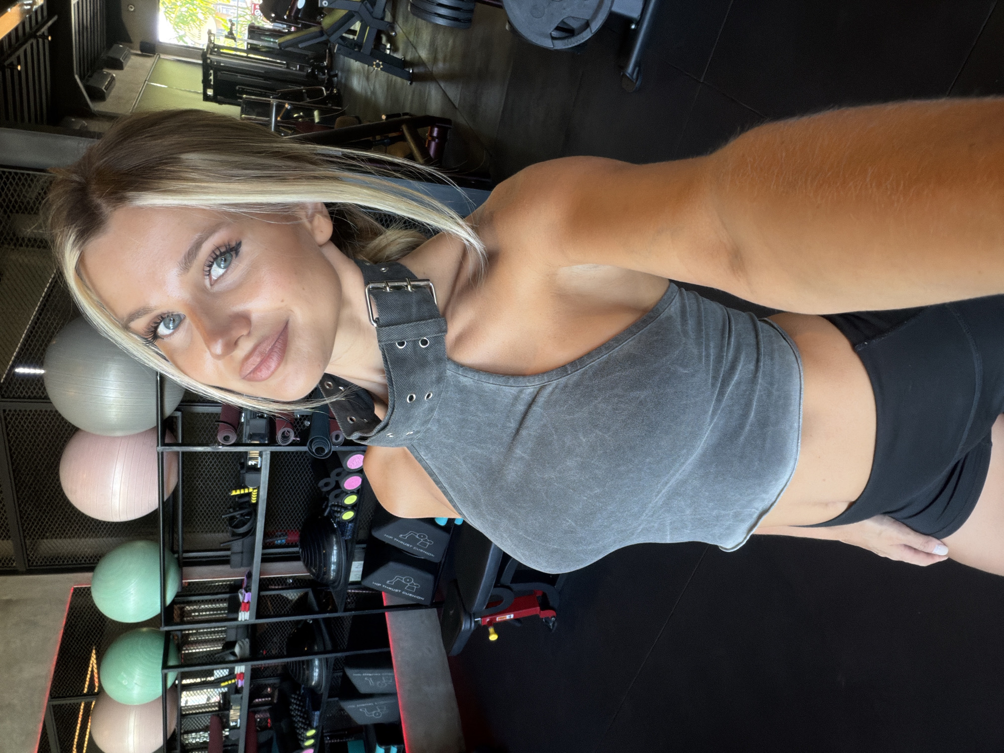 In a brightly lit gym located in North Kuta, Indonesia, a young woman with blonde hair and blue eyes is taking a selfie. She is wearing a grey, cut-out crop top and black athletic shorts. Behind her, gym equipment is visible, including weight machines, exercise balls, and mats. The atmosphere appears energetic and focused on fitness.