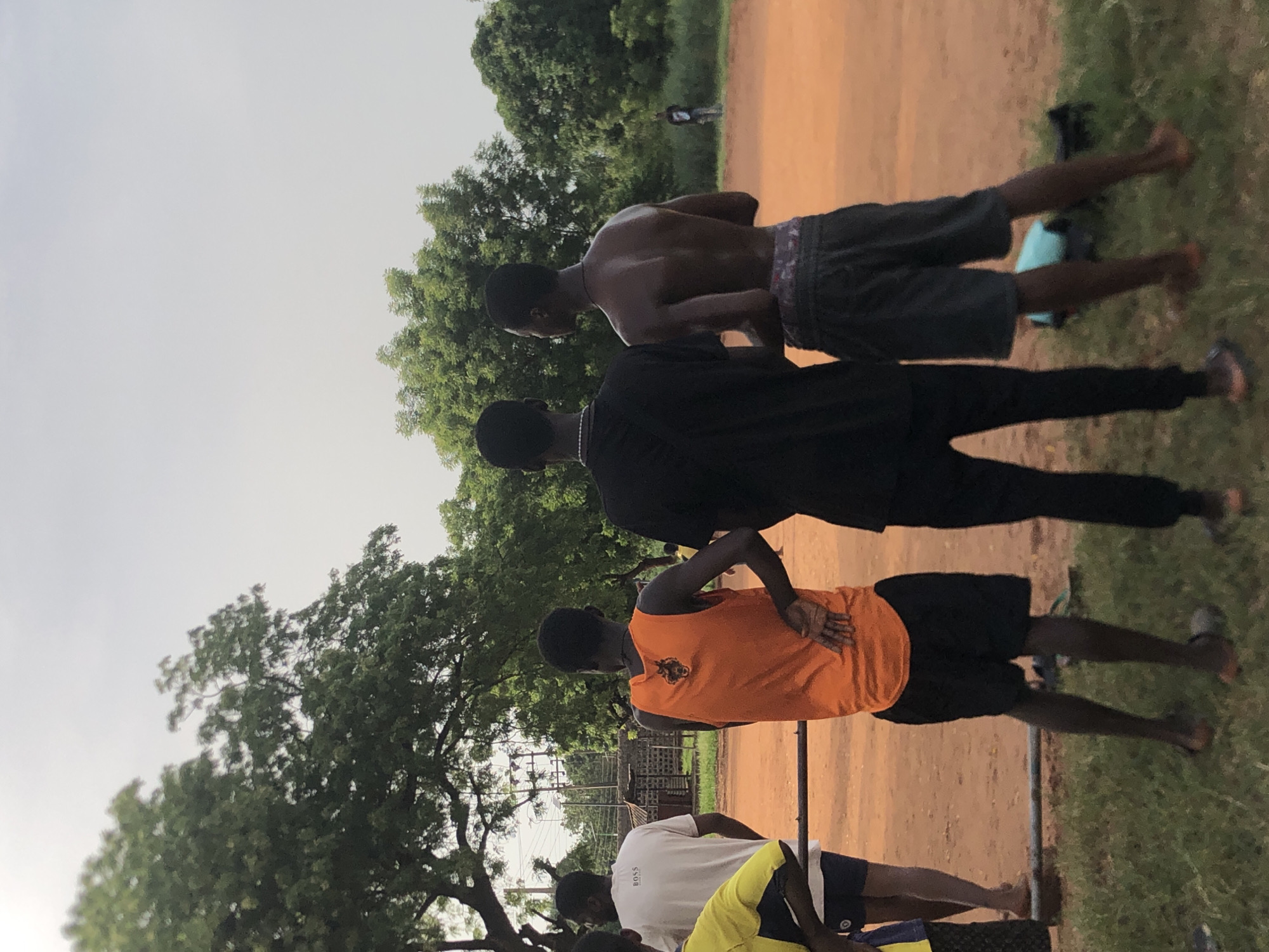 The image depicts a group of young men gathered outdoors on a reddish-brown dirt path, characteristic of a community field or open ground, in Ukwu-Abwa, Nigeria. The scene is illuminated by diffused daylight, suggesting an overcast sky or the soft light of early morning or late afternoon. Lush green trees with dense foliage dominate the background and extend into the left side of the foreground.
In the mid-ground, three men are seen from behind. The man on the right is shirtless, wearing dark shorts with a patterned waistband. Next to him, a man in a black long-sleeved shirt and black trousers stands. To his left, another man wears a bright orange sleeveless vest featuring a dark logo or design on the back, paired with black shorts; his hands are placed on his lower back, possibly indicating a stretch or relaxed pose.
In the foreground, partially visible, are additional individuals. One wears a white T-shirt with the word "BOSS" clearly printed on the back, alongside dark shorts and a yellow garment. A slender metal bar or fence runs horizontally across the lower part of the frame. In the distant background, beyond the trees, a faint figure and what appear to be utility poles or power lines can be discerned. The ground consists mainly of dirt but includes patches of green grass, particularly on the right side and in the immediate foreground.
The men appear to be casually standing or waiting, suggesting a relaxed social gathering or preparation for an outdoor activity.
