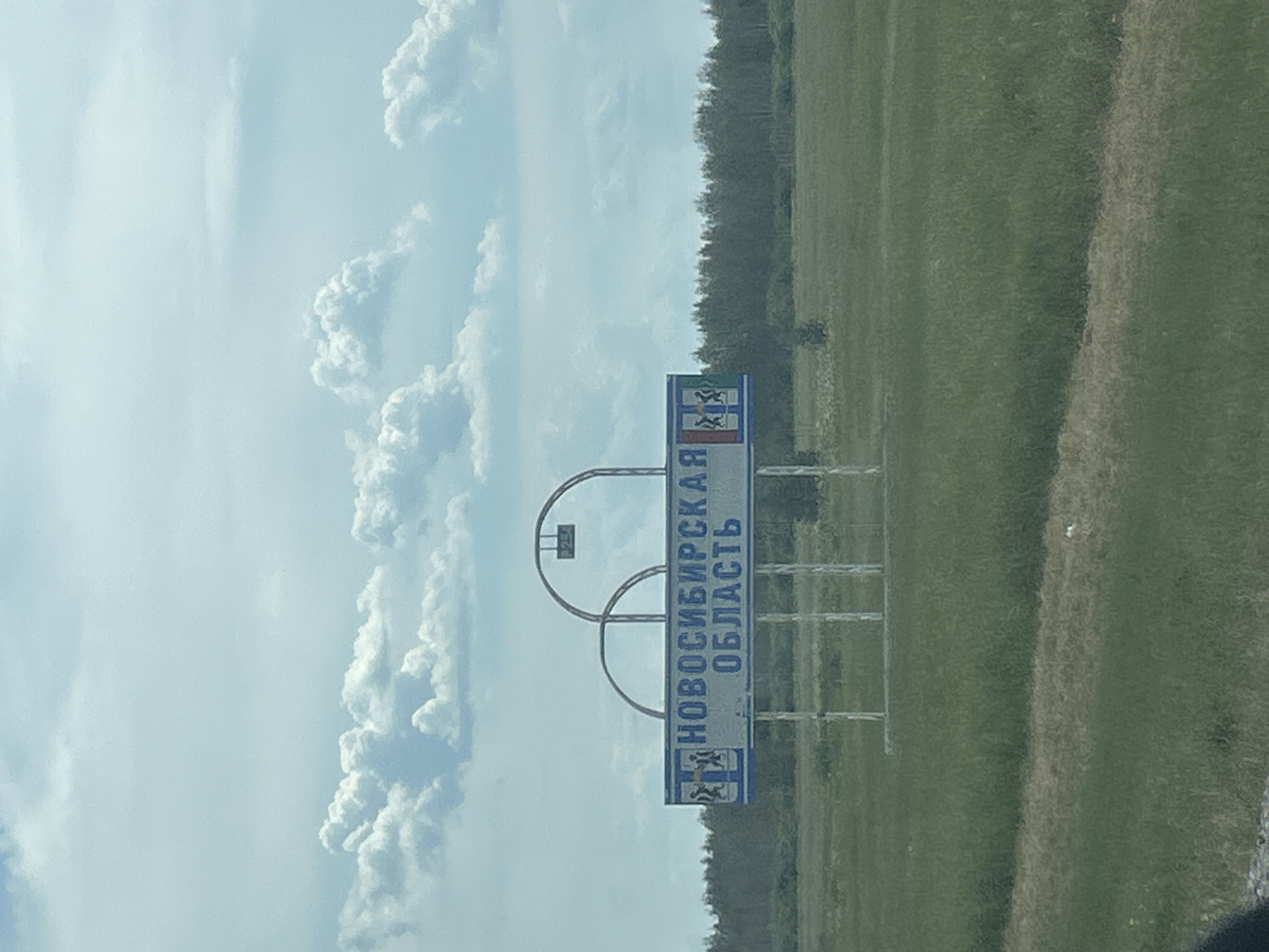 The image captures a roadside sign indicating the Novosibirsk Oblast in Russia. The sign is white with blue lettering and features a coat of arms on the left side. The text on the sign reads "НОВОСИБИРСКАЯ ОБЛАСТЬ". The sign is supported by metal poles and is positioned in a rural landscape with green fields and a distant treeline under a cloudy sky. There are no people visible in the image.