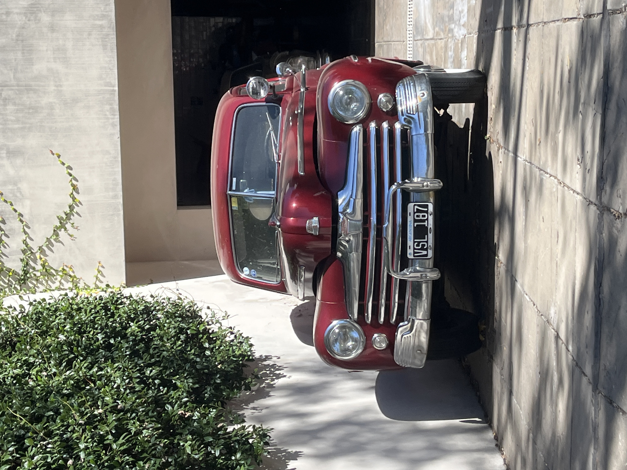 A vintage, deep red fire truck is the central subject, parked outdoors on a bright, sunny day. The vehicle features a prominent chrome grille with vertical bars, large circular headlights, and smaller auxiliary lights positioned below. A chrome bumper is visible at the front, beneath which is a license plate displaying "ARGENTINA" at the top and "VSL 187" as the registration number. The driver's side door and window are visible, along with a small, round chrome mirror. A spare tire is mounted on the side of the truck.
The truck is situated on a light-colored, paved surface, which is marked by strong, distinct shadows, indicating bright midday sunlight. To the left of the vehicle, a dense, dark green bush or hedge occupies the foreground, with climbing ivy visible on a light-colored wall behind it. To the right and behind the truck, a darker wall or entrance suggests a building or garage. The overall scene depicts a clear, warm day in Olivos, Argentina, with the vintage vehicle as the focal point.