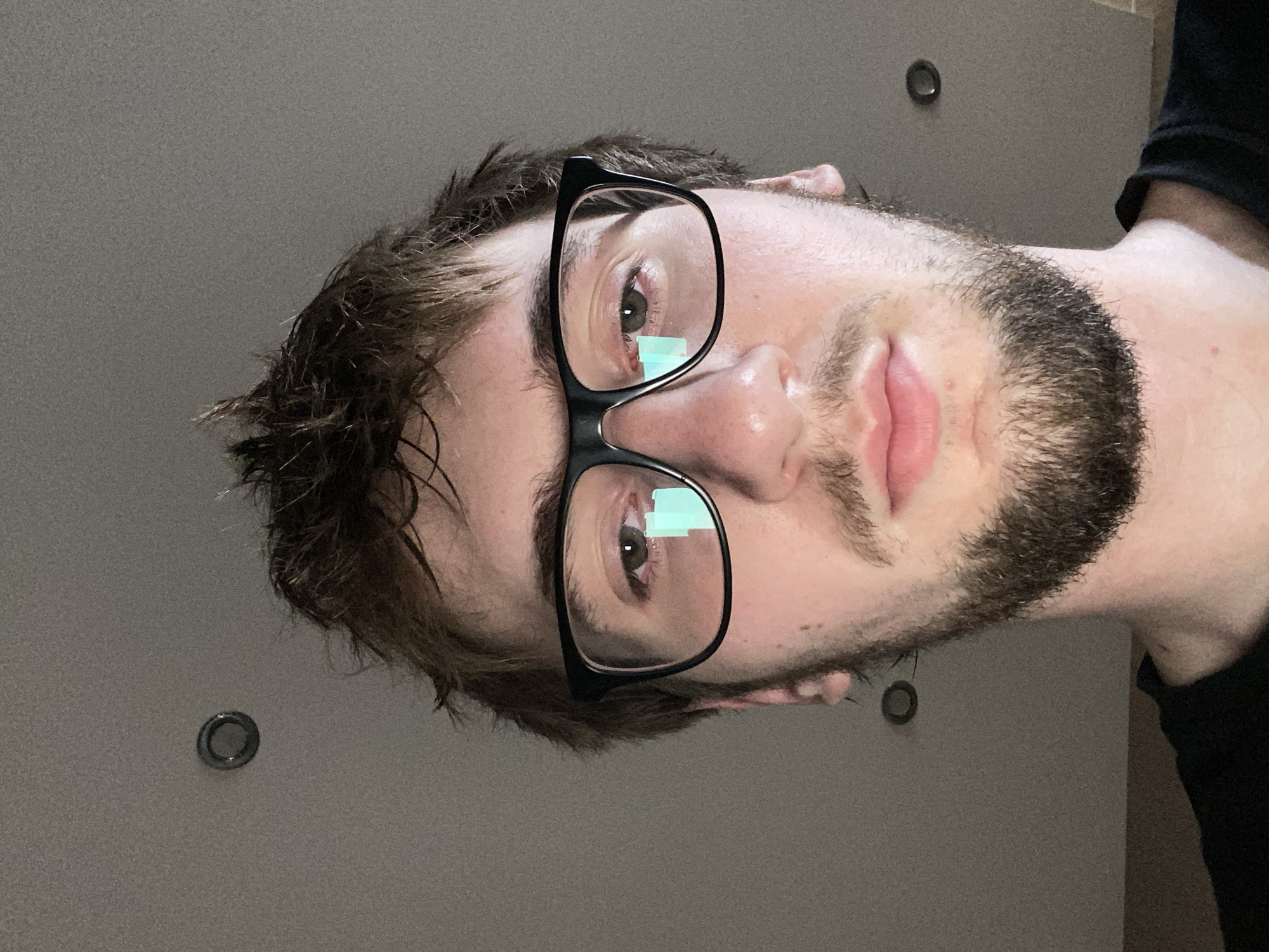 The image is a close-up, low-angle selfie of a young man wearing black-rimmed glasses and a black t-shirt. He has light brown, messy hair and a short beard. His eyes are visible through the glasses, with a reflection of what appears to be a blue screen on the left lens. The background is a ceiling with two recessed lights, suggesting an indoor environment. There is no information available in the image to determine the location in the city of Khmelnytskyi, Ukraine.