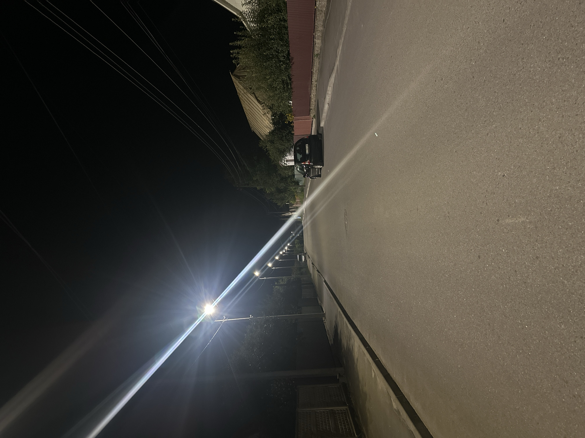 The image depicts a street scene at night in Khmilnyk, Ukraine, likely in a residential or suburban area. The street is asphalted and appears to be a two-way road. Several streetlights illuminate the scene, casting long streaks of light due to the long exposure or aperture setting. On the left side of the road, there is a sidewalk and a dark fence, possibly made of wood or metal, with a patterned design. Trees and foliage line the side of the street behind the fence. On the right side, houses with pitched roofs and a reddish-brown fence are visible. A dark-colored car, possibly a hatchback, is parked on the right side of the road. Power lines are strung across the dark night sky above the street. The overall impression is a quiet, illuminated street at night.