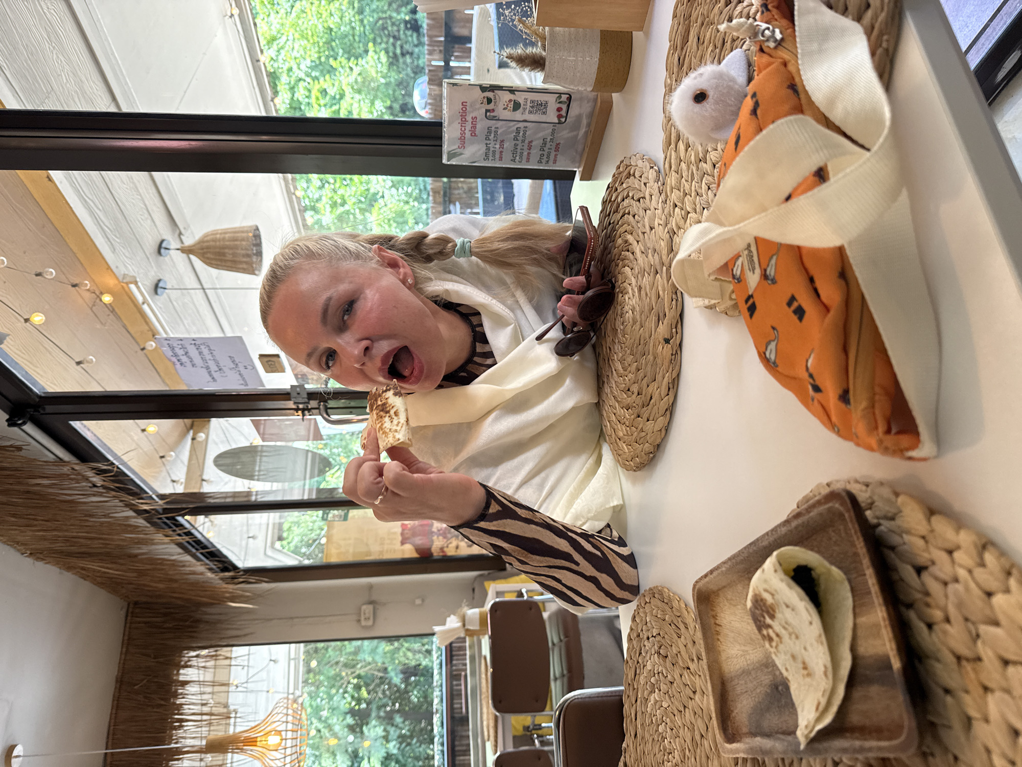 A blonde woman with her hair in two braids is sitting at a white table in a cafe in Koh Samui, Thailand. She is wearing a white shawl over a patterned top and is about to eat a piece of flatbread. In front of her is a small wooden dish with a folded flatbread filled with a dark filling. To her right is a brochure advertising subscription plans, sunglasses, and a smartphone. A white plush toy with large eyes is placed on an orange fabric bag in the foreground. The cafe has a rustic decor with thatched accents and woven placemats. Large windows offer a view of lush greenery outside.