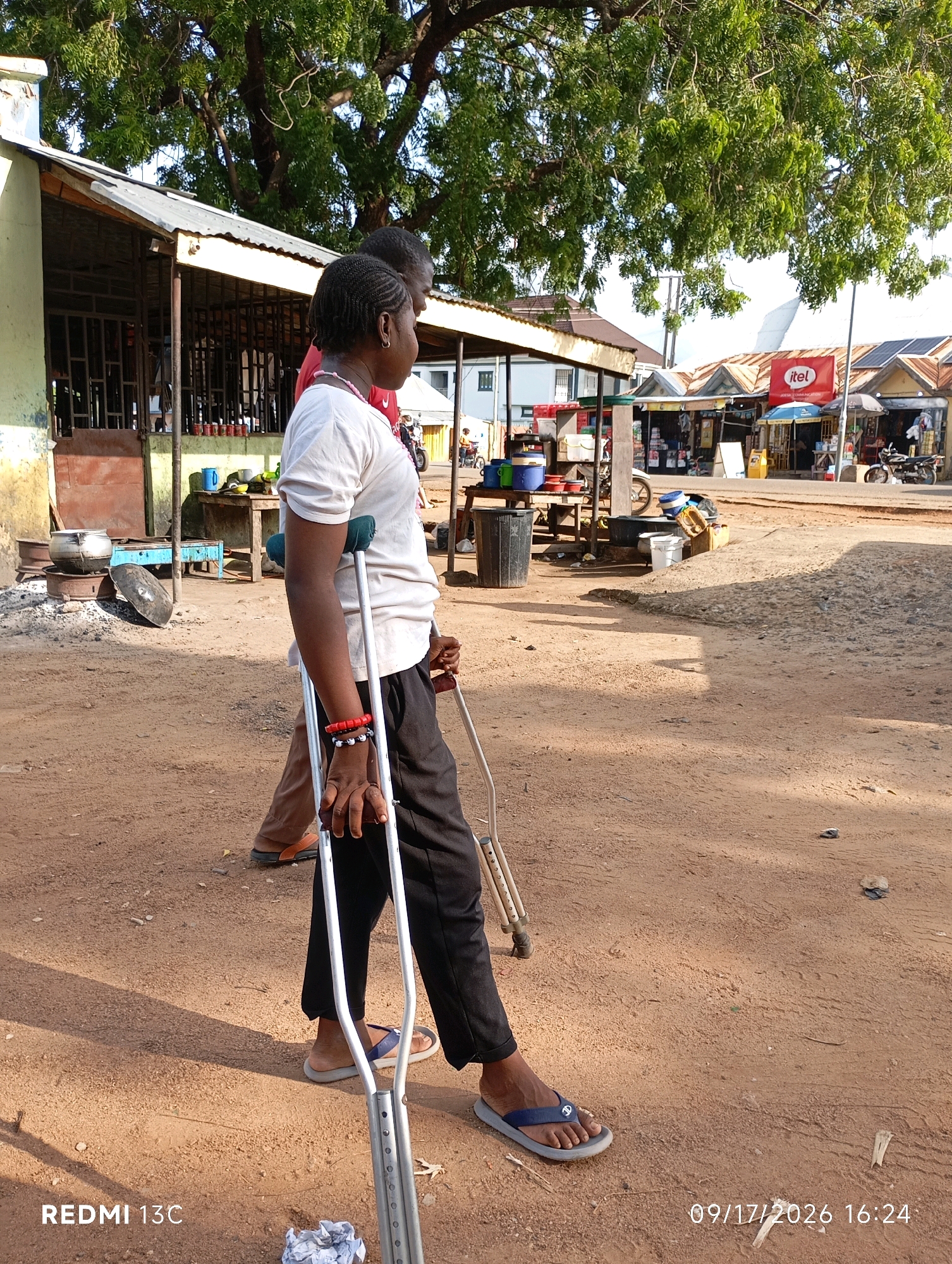 In the city of Shendam, Nigeria, a woman with braided hair is walking using forearm crutches. She is wearing a white t-shirt, black pants, and blue flip-flops. She has red and white bracelets on her left wrist. Beside her, a person in brown pants and orange sandals walks in the same direction. The ground is covered in reddish-brown dirt, and there are scattered pieces of debris. In the background, there are several structures that appear to be market stalls or shops with corrugated metal roofs. One stall displays a red sign with the "itel" logo. Further in the background, a tree with lush green foliage provides shade. Sunlight casts distinct shadows on the ground.