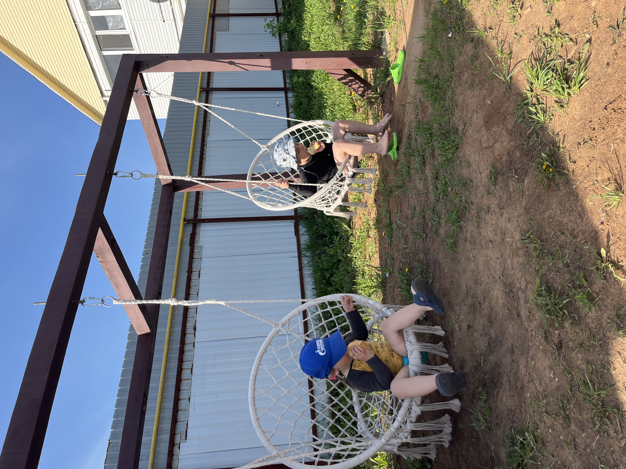 Two children sit in hanging macrame chairs suspended from a dark brown wooden frame. One child wears a blue baseball cap, yellow and black shirt, and blue swimming trunks; they are wearing blue flip-flops. The second child is wearing a patterned hat, a black long-sleeved shirt, and green flip-flops. The location is in a yard in the city of Chaikovsky, Russia, with a metal fence visible in the background. The ground is dirt mixed with patches of green grass. A house with white siding is visible in the upper left corner.