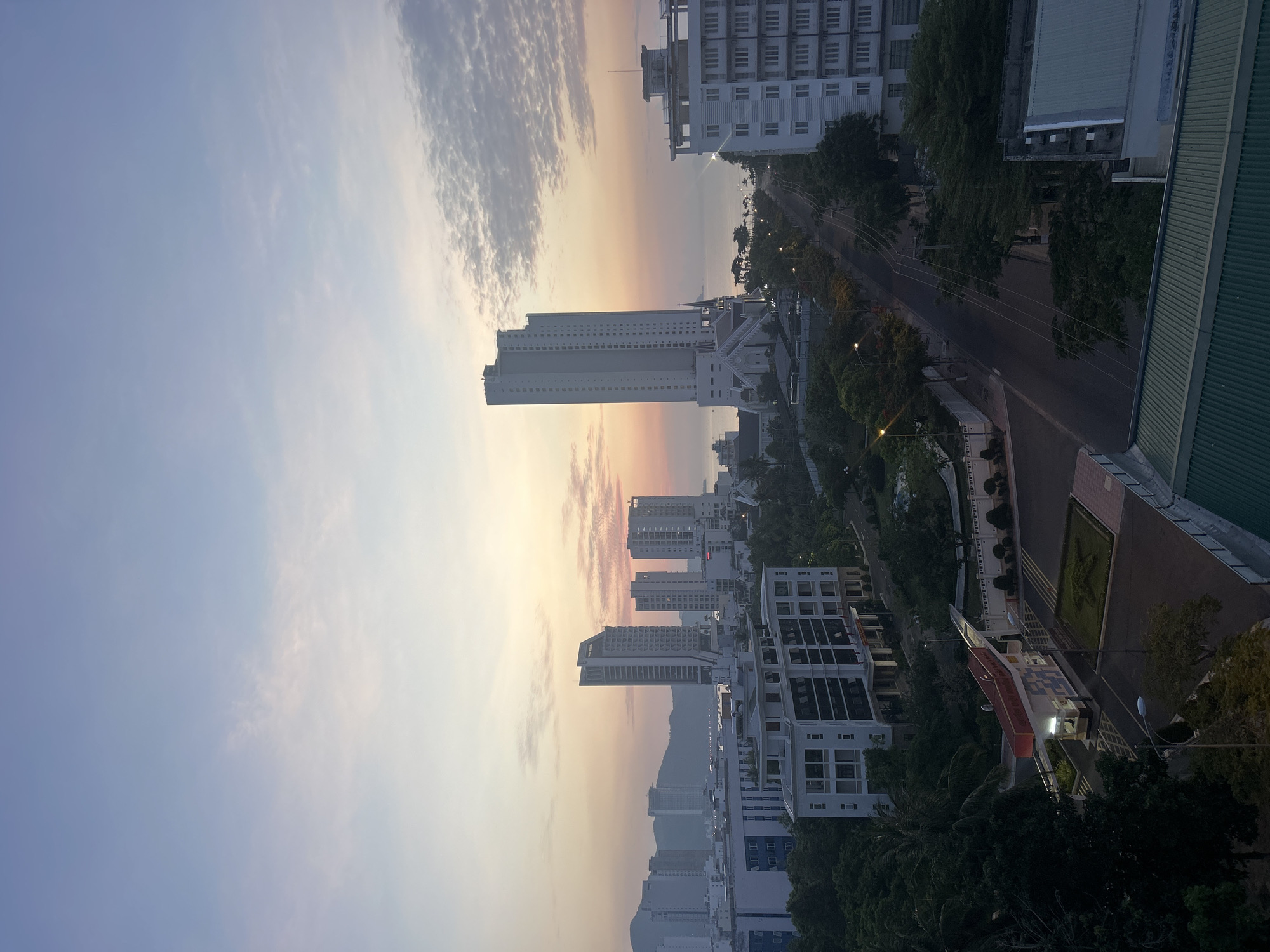 The image shows an urban landscape at sunset in Bac Nha Trang Ward, Vietnam. The sky transitions from blue to orange near the horizon, with some cloud cover. Several high-rise buildings dominate the skyline, and other structures are visible, including a building with a green roof. A road runs through the scene lined with trees. Streetlights illuminate the area. The image appears to be taken from an elevated vantage point.