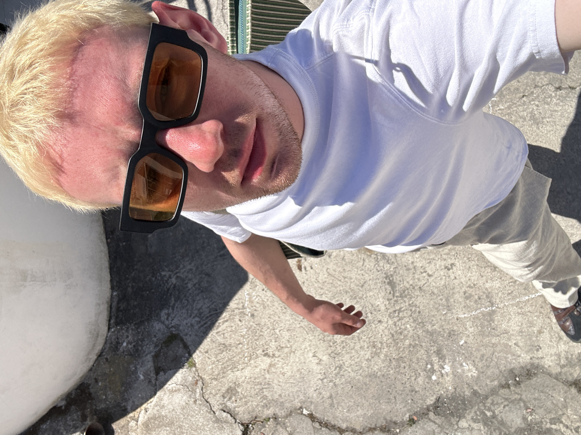 This overhead, high-angle shot features a young adult male standing outdoors in Lipari, Italy, under strong daylight. He is looking upwards towards the camera, his face partially obscured by modern black sunglasses with amber-tinted lenses. The man has short, bleached blonde hair and light stubble on his chin and jawline. He is wearing a white crew-neck t-shirt and light-colored, possibly beige or light grey, trousers.
The bright sunlight casts sharp, well-defined shadows on the concrete ground, indicating a clear, sunny day. The ground is a textured light grey concrete with visible cracks. To the left, a large, smooth, light-colored wall or structure is prominent. The upper right portion of the image shows a hint of a striped pattern, possibly part of an awning or outdoor furniture. The bottom right corner reveals the toes of dark, possibly brown, shoes. The setting suggests a casual, warm outdoor environment. There is no visible text within the image.