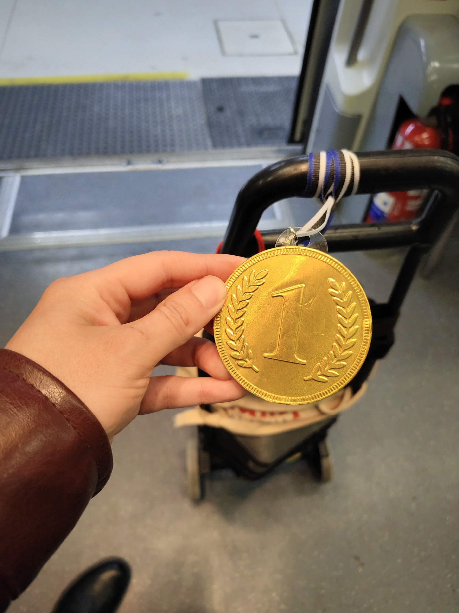 A first-person view photograph shows a hand holding a gold medal with the number "1" prominently displayed, surrounded by laurel wreaths. The medal hangs from a black trolley with blue and white striped straps, likely used for carrying items. The trolley seems to be inside a vehicle with a floor covered in gray and black non-slip matting, possibly a bus or van. A red fire extinguisher is visible in the background, mounted on the side wall of the vehicle. A leather-sleeved arm suggests the person holding the medal is wearing a coat. The background suggests an industrial or public transport setting. The photograph does not contain any information pertaining to the location of the city of Barcelona, Spain.