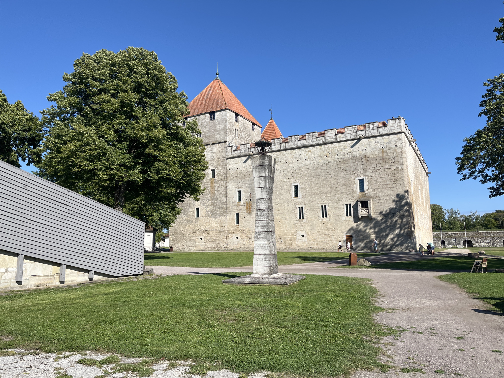 In the city of Kuressaare, Estonia, a large, weathered stone castle with red-tiled roofs stands under a clear blue sky. A tall, segmented stone monument is situated in the foreground on a grassy area. Adjacent to the monument, a path curves through the grass. To the left, a modern, grey, sloped wooden structure partially obscures the view. Several people are visible in the background near the castle, walking and interacting. The scene is bathed in sunlight, casting shadows across the grounds. The castle appears to be a historical landmark, with its stone walls and arched windows. The overall setting suggests a public park or historical site, with lush green grass and trees contributing to the natural environment.