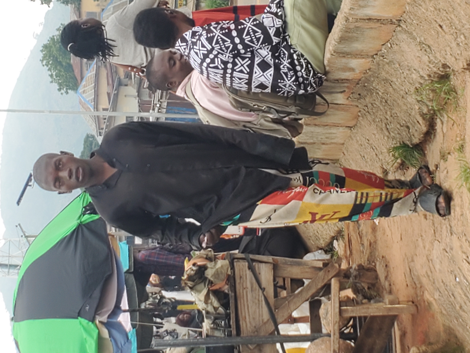 In Gwong, Nigeria, a young man stands in the foreground wearing a black collared shirt and vibrantly patterned trousers. His right leg is forward, and he is wearing black sandals. Behind him, a market stall with a green and black umbrella is visible, piled with various goods. Further in the background, several people are seated on a low wall, including a woman with her back to the camera, and two men. One man wears a pink shirt and a backpack, while the other has a black and white patterned shirt and is looking towards the left. The environment appears to be an outdoor market setting with a dirt path and a stone wall.