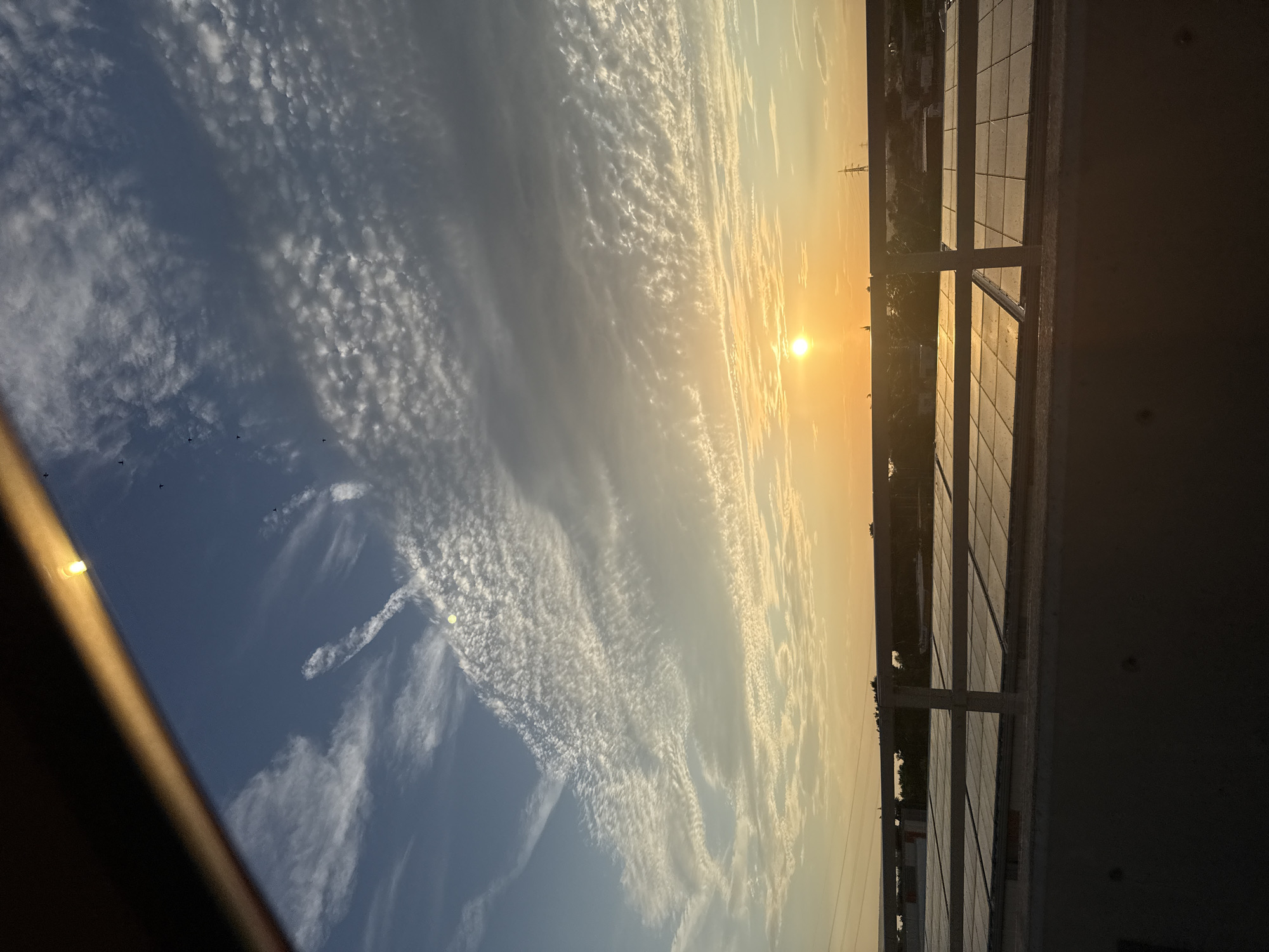 The image captures a panoramic view of a sunrise over an urban landscape, likely Burgas, Bulgaria, given the provided context. The sun, a bright orb of golden light, is low on the horizon, casting a warm glow across the sky. Wispy clouds, tinged with hues of orange and yellow, stretch across the blue expanse, creating a textured and dynamic sky.
In the foreground, a balcony railing with a tiled floor suggests a vantage point from a building, possibly a hotel or residential apartment. The railing is made of metal and has a modern design, with vertical and horizontal bars. Beyond the railing, the scene unfolds: a distant cityscape with buildings and infrastructure, including what appear to be power lines and possibly some industrial structures. Trees and greenery are visible, dotting the landscape between the buildings.
The overall atmosphere is serene and tranquil, evoked by the soft light and the expansive sky. The time of day is clearly dawn, indicated by the rising sun and the gentle, warm illumination. The weather appears clear, with the clouds adding visual interest to the sky rather than suggesting inclement conditions. A few small, dark shapes, possibly birds, are visible in the sky, adding a sense of scale and life to the scene. The image has a slightly tilted perspective, as if the camera was held at an angle. No text is visible in the image.