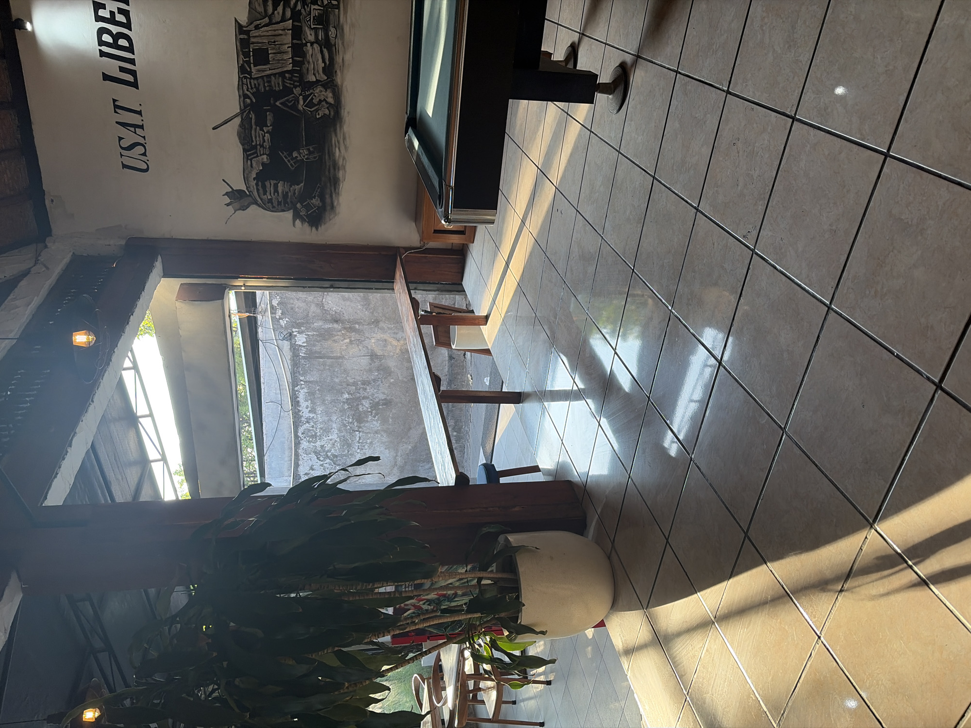 This is a photograph taken from a ground-level perspective inside what appears to be an indoor-outdoor space, possibly a cafe or recreational area, in Kubu, Indonesia. The scene is dominated by natural light streaming in from an opening to the right, casting long shadows across the tiled floor. In the foreground, a large potted plant with dark green leaves stands near a wooden pillar. To the left, a few wooden chairs and tables are visible, suggesting a seating area.
Further into the space, a long, narrow wooden table is mounted on the wall, with a small blue stool placed underneath it. Beyond this, a grey concrete wall is visible. The right side of the image features a pool table, with its green felt surface reflecting the sunlight. On the white wall behind the pool table, a black mural is depicted, showing a tank and what appears to be a word in black lettering above it: "USAT LIBER". The floor is covered in light-colored square tiles. The overall impression is one of a casual, sunlit establishment with recreational amenities.