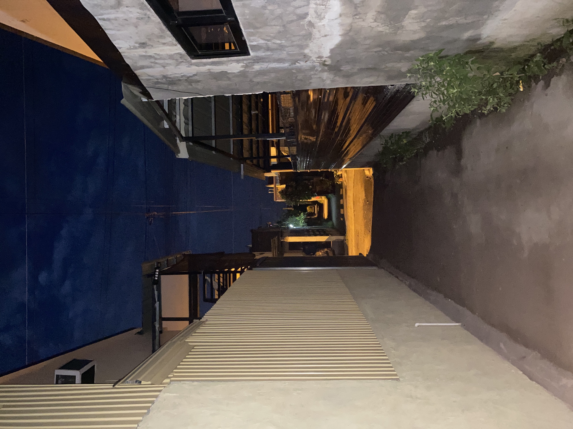 The image depicts a narrow alleyway at dusk in the city of Davao City, Philippines. The alley is flanked by two structures. On the left, a building with a textured, light-colored wall and a beige slatted fence dominates the foreground. An air conditioning unit is visible on the upper part of this building. On the right, a rough, concrete wall with a dark framed window is prominent. This wall is adjacent to a structure with a corrugated metal roof supported by dark metal beams, with some reflective material visible underneath. Overgrown green plants are growing along the base of the concrete wall on the right.
The alleyway itself is paved with a dark, muted surface, likely asphalt or concrete, and is illuminated by artificial light sources that cast warm yellow glows in the distance. The sky above is a deep blue, transitioning from twilight hues near the horizon, with some wispy clouds visible. Electrical wires are strung across the sky. The overall impression is one of a residential or possibly commercial back alleyway in an urban setting, viewed from a low angle. There are no people or animals visible in the immediate scene. The scene suggests a quiet, possibly late evening or early morning ambiance.