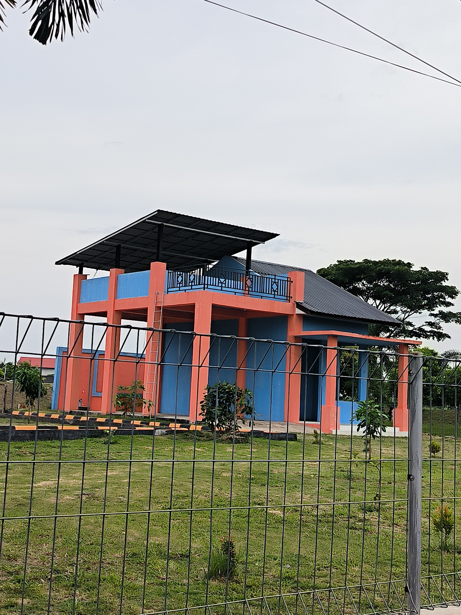 A two-story building with orange and blue walls stands behind a wire fence. The building has a dark gray roof and an open second story with a black railing. A ladder leans against the side of the building. The surrounding area is grassy with some small trees and a larger tree in the background. The sky is overcast. The building is located in the city of Lhokseumawe, Indonesia.