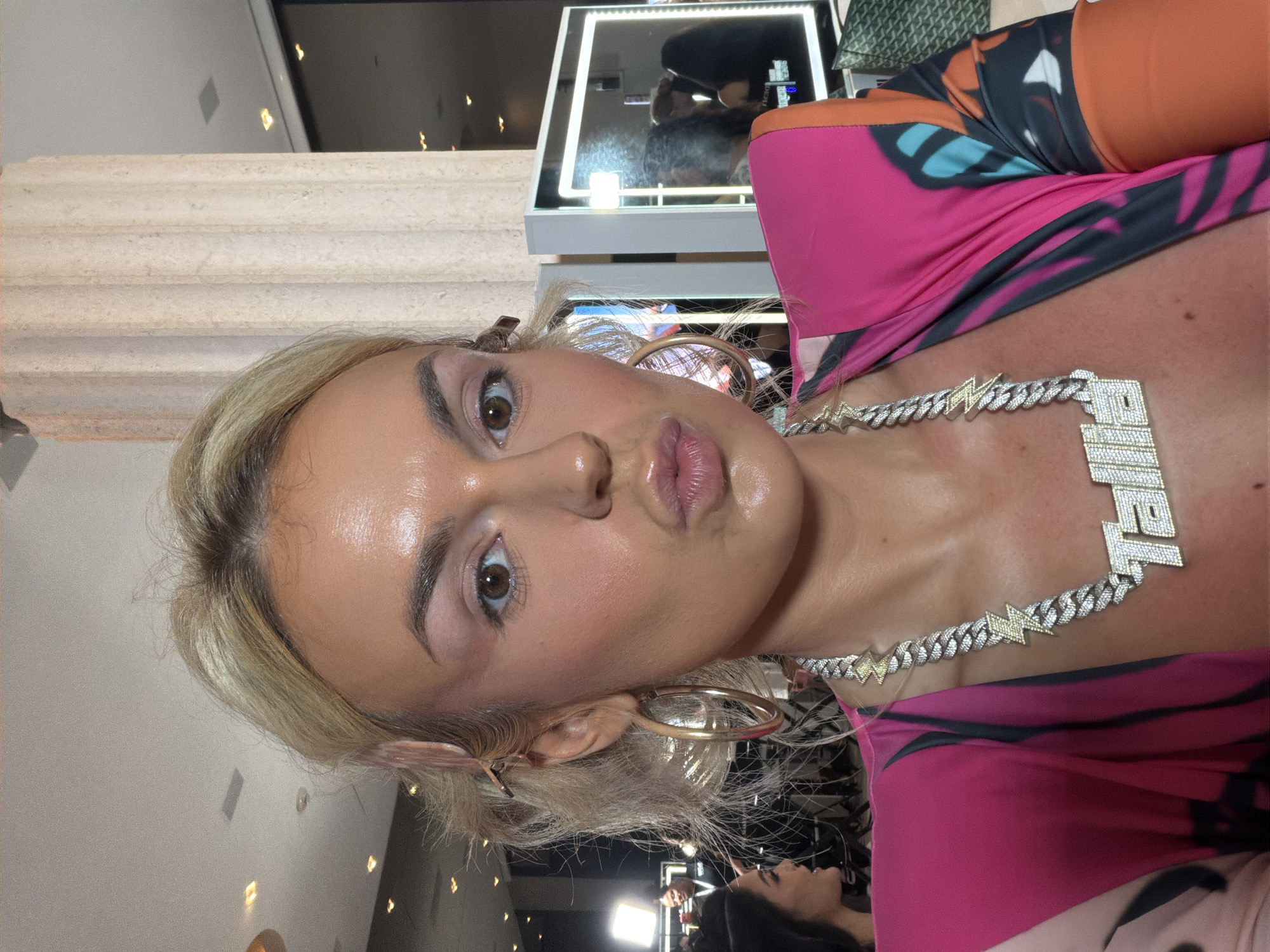 A medium close-up shot shows a blonde woman with light skin in Miami Beach, USA. She has brown eyes, full lips, and wears large gold hoop earrings. She also wears a large, diamond-encrusted necklace with the name "TRIEL" spelled out. She is wearing a pink and black top, and her face is shiny. In the background, there is a person in the lower center frame and a display case with a reflection in the upper center frame. The background is blurred. There is also a beige pillar on the left.