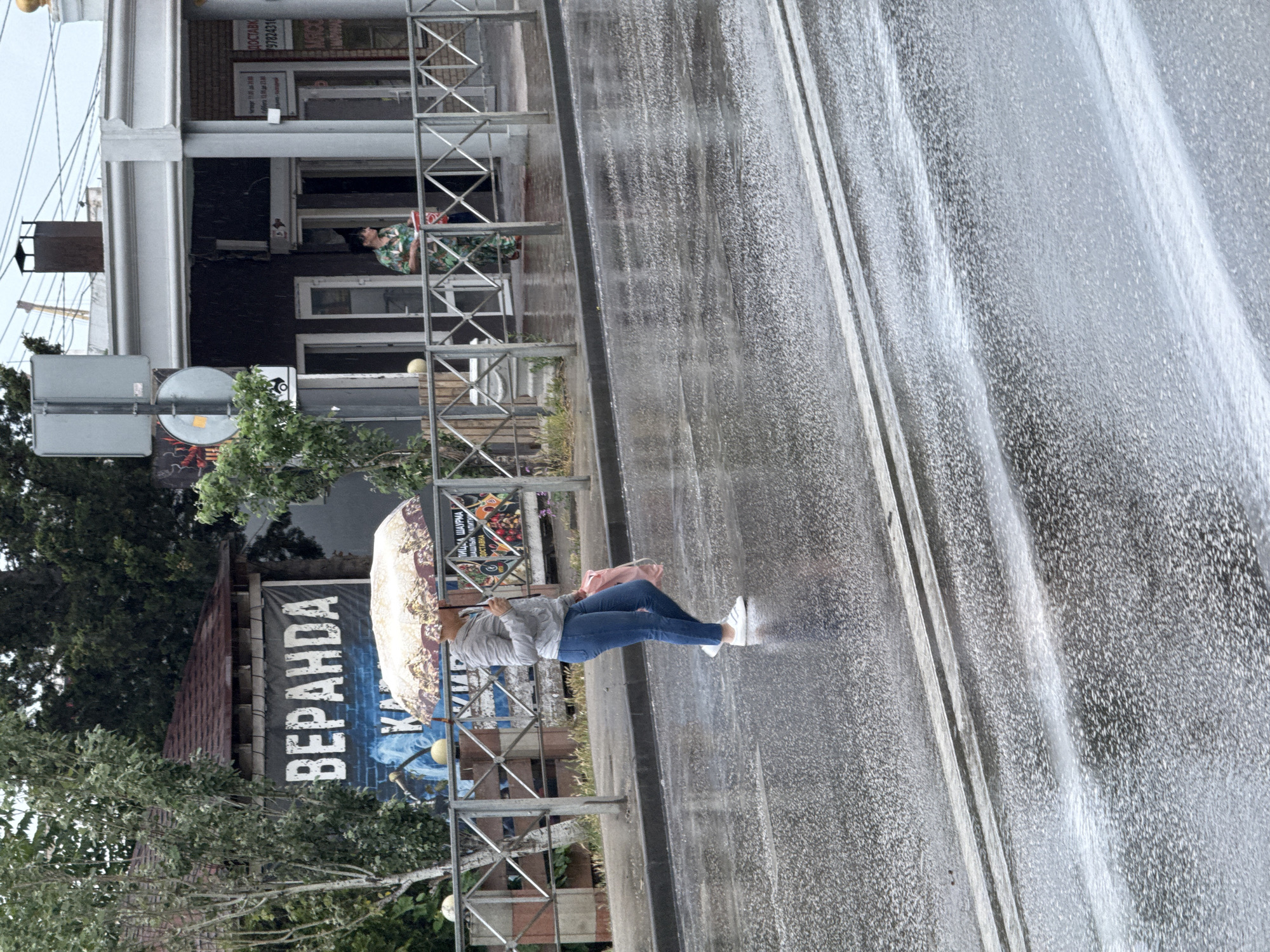 A woman with an umbrella walks on a wet street in an urban environment. The woman is wearing a gray hooded jacket, blue jeans, and white shoes. She is holding a patterned umbrella over her head and a pink bag in her right hand. The street is wet and reflects the light, indicating it is raining or has recently rained. In the background, there are buildings, trees, and a metal fence. One building has a sign with the word "ВЕРАНДА" (Veranda) in large white letters. Another building has signs and an open doorway. Another person in a green and black patterned shirt is standing near the doorway of a building. The sky appears overcast.