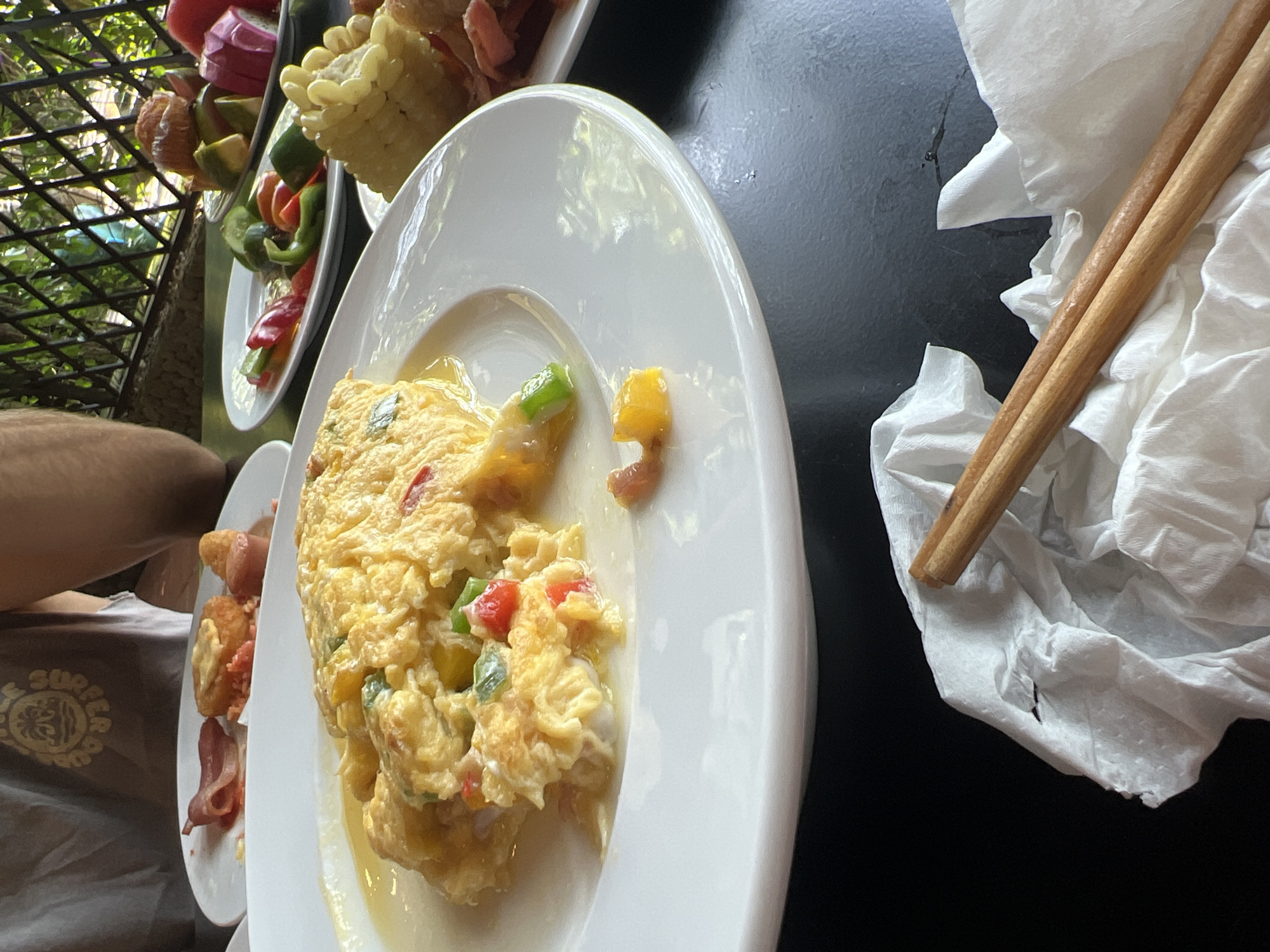The image depicts an outdoor meal, likely breakfast or brunch, served on a bright day. In the foreground, a white plate holds a serving of scrambled eggs or an omelette, mixed with diced red and green bell peppers and possibly some corn kernels. A small amount of liquid, likely egg drippings, is visible at the bottom of the plate.
To the immediate left of this plate, another white plate is partially visible, containing strips of bacon and what appears to be a fried potato item, such as a hash brown. Further to the left, the lower arm and leg of a person are visible. The person is wearing a brown or grey t-shirt with a circular logo that clearly reads "SURF HOUSE" above a wave design, suggesting a relaxed, beach-oriented style.
In the upper left background, behind the plates, colorful food skewers are visible, featuring tropical fruits like red dragon fruit, yellow mango, and possibly watermelon, alongside green and red bell peppers and small pieces of corn on the cob. The background also shows lush green foliage and a dark metal lattice or railing, indicating an outdoor dining setting, possibly on a patio or balcony.
On the dark table surface to the right of the main plate, a crumpled white napkin holds a pair of wooden chopsticks. The table surface is dark, possibly black, with visible reflections from the bright natural light. The overall scene, with tropical fruits, an outdoor setting, and the use of chopsticks, is consistent with the provided location context of Đà Nẵng, Vietnam, and suggests a pleasant daytime meal.